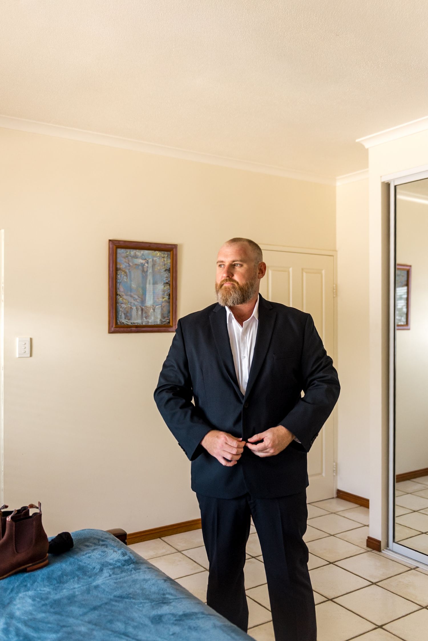 Bearded groom in dark navy suit buttoning jacket while getting ready for wedding ceremony indoors.