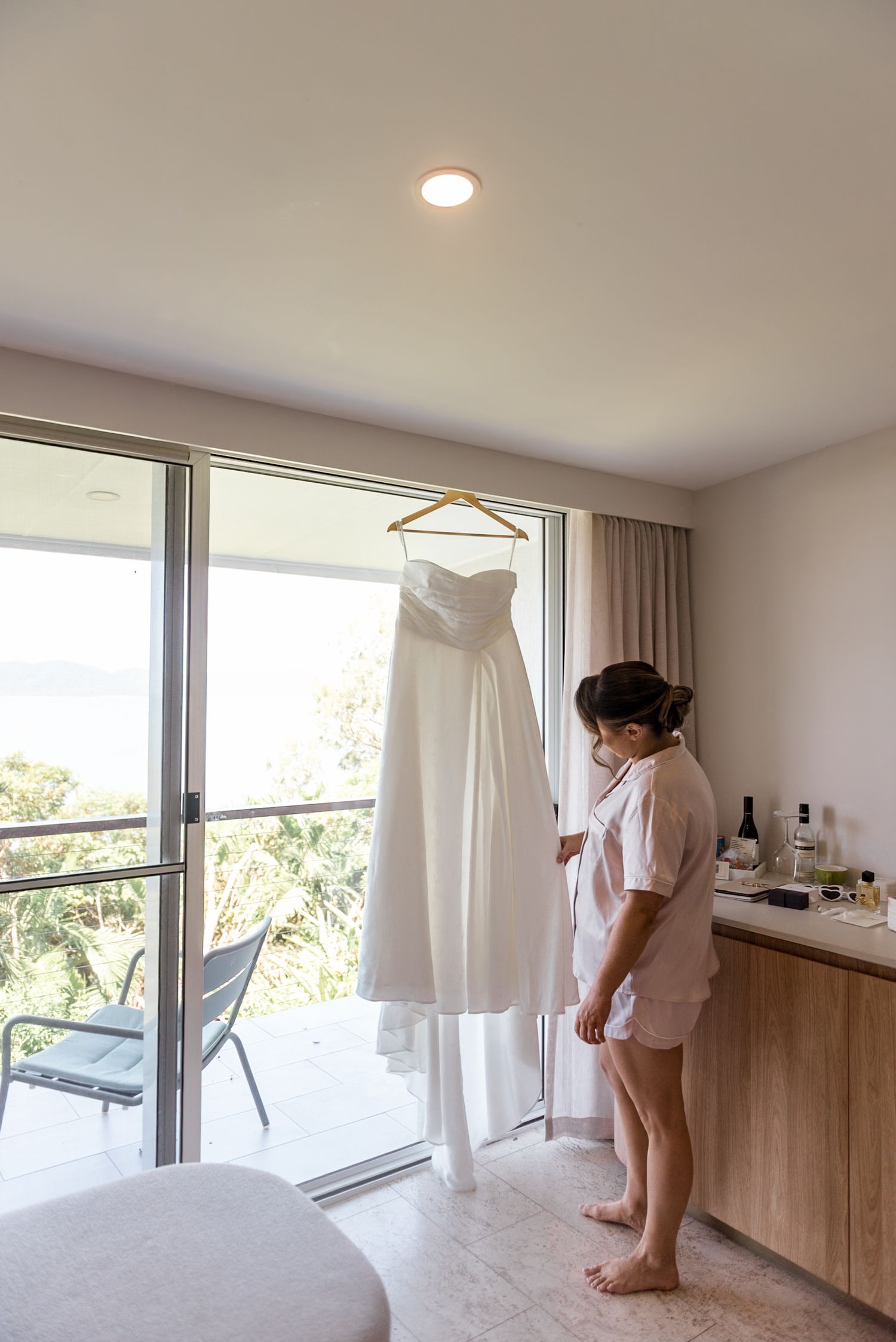 Bride in pink robe admires her strapless white wedding gown hanging by a window with ocean view.