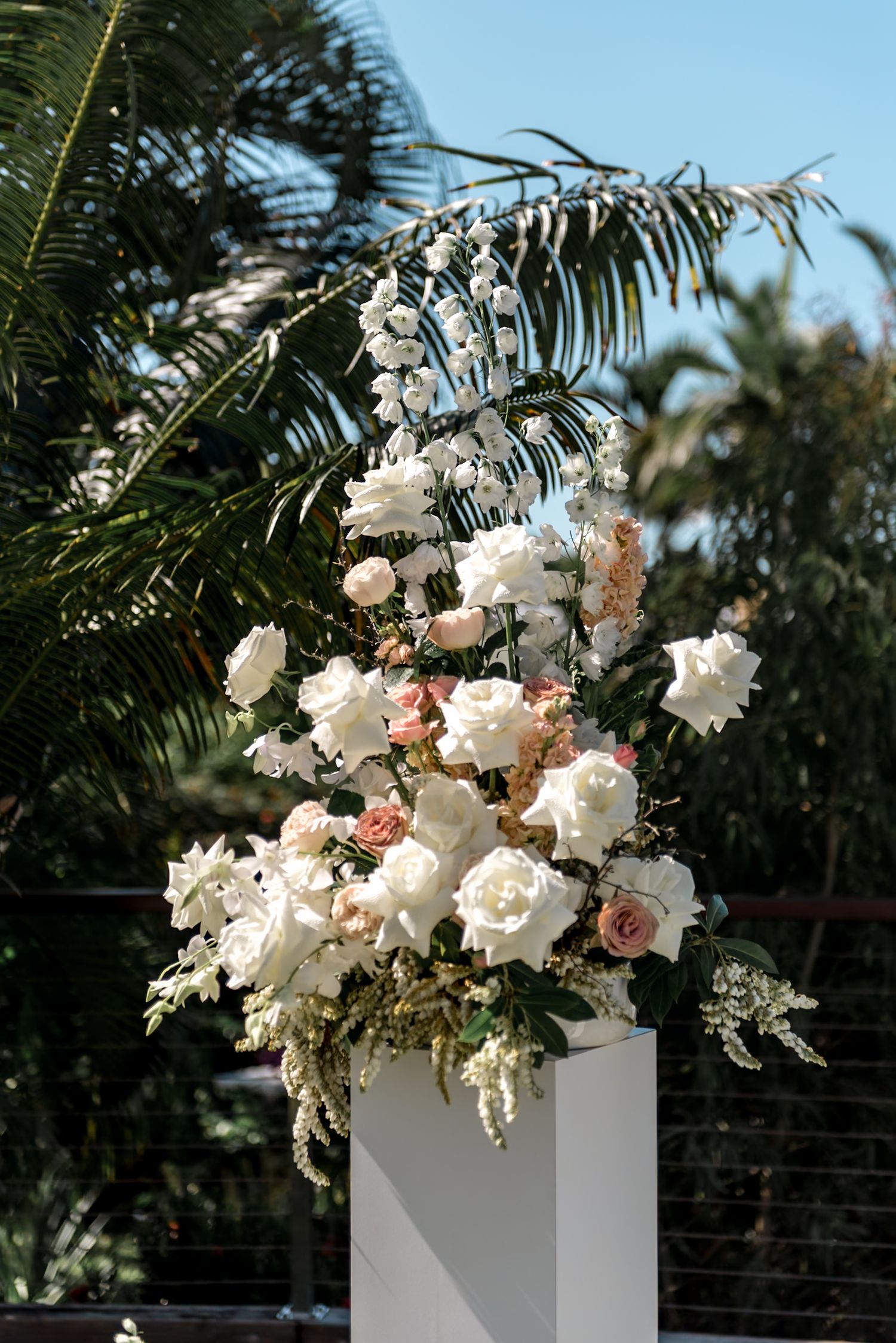 Close-up of a stunning white and blush rose floral arrangement on a white pedestal with tropical palm trees behind.