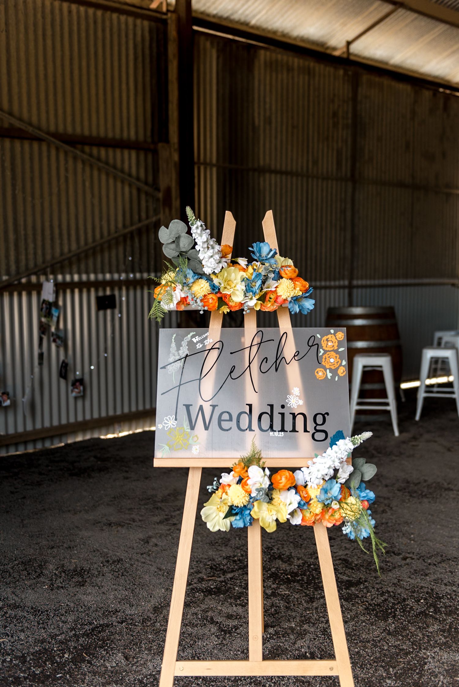 Fletcher Wedding welcome sign on easel decorated with blue, orange, and yellow flowers in a barn.