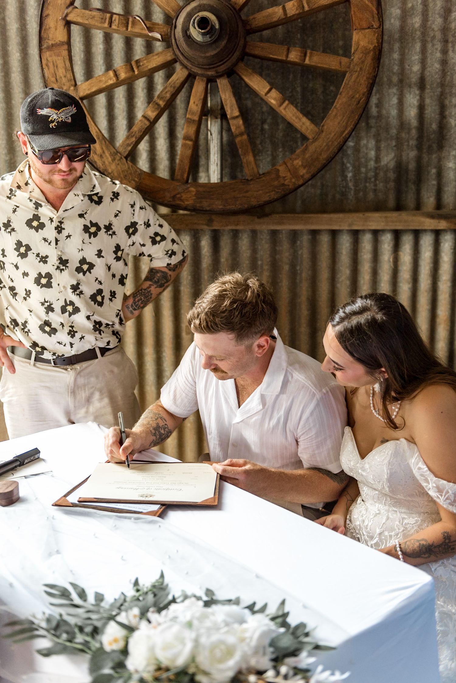 Newlyweds signing marriage certificate in rustic barn with witness looking on.