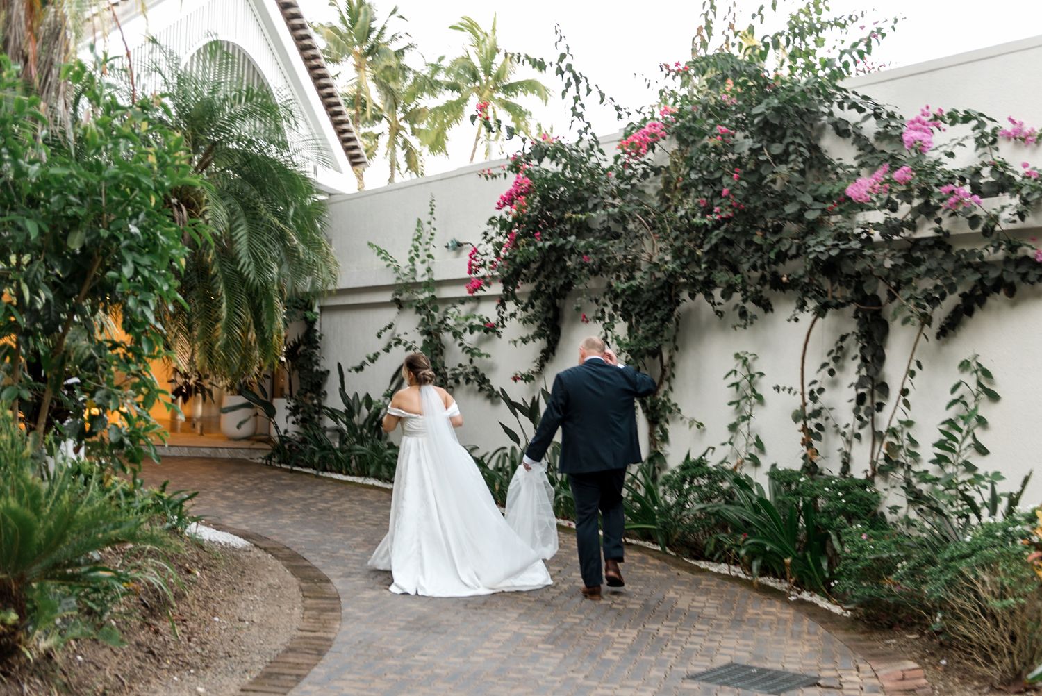 Bride and groom walking through a lush tropical garden pathway on their wedding day.