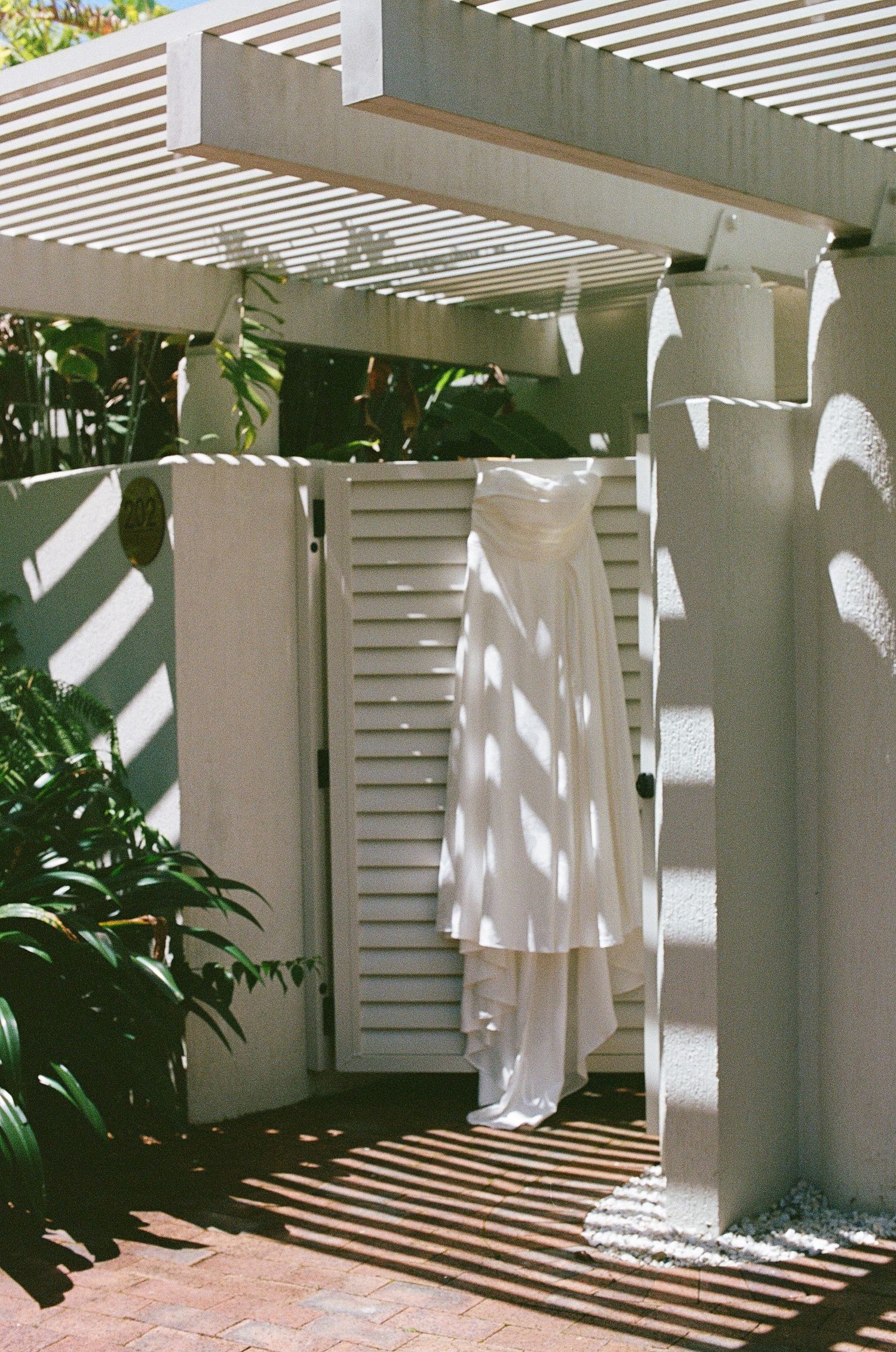 Elegant white wedding dress hangs on pergola gate casting beautiful shadows in sunny courtyard.