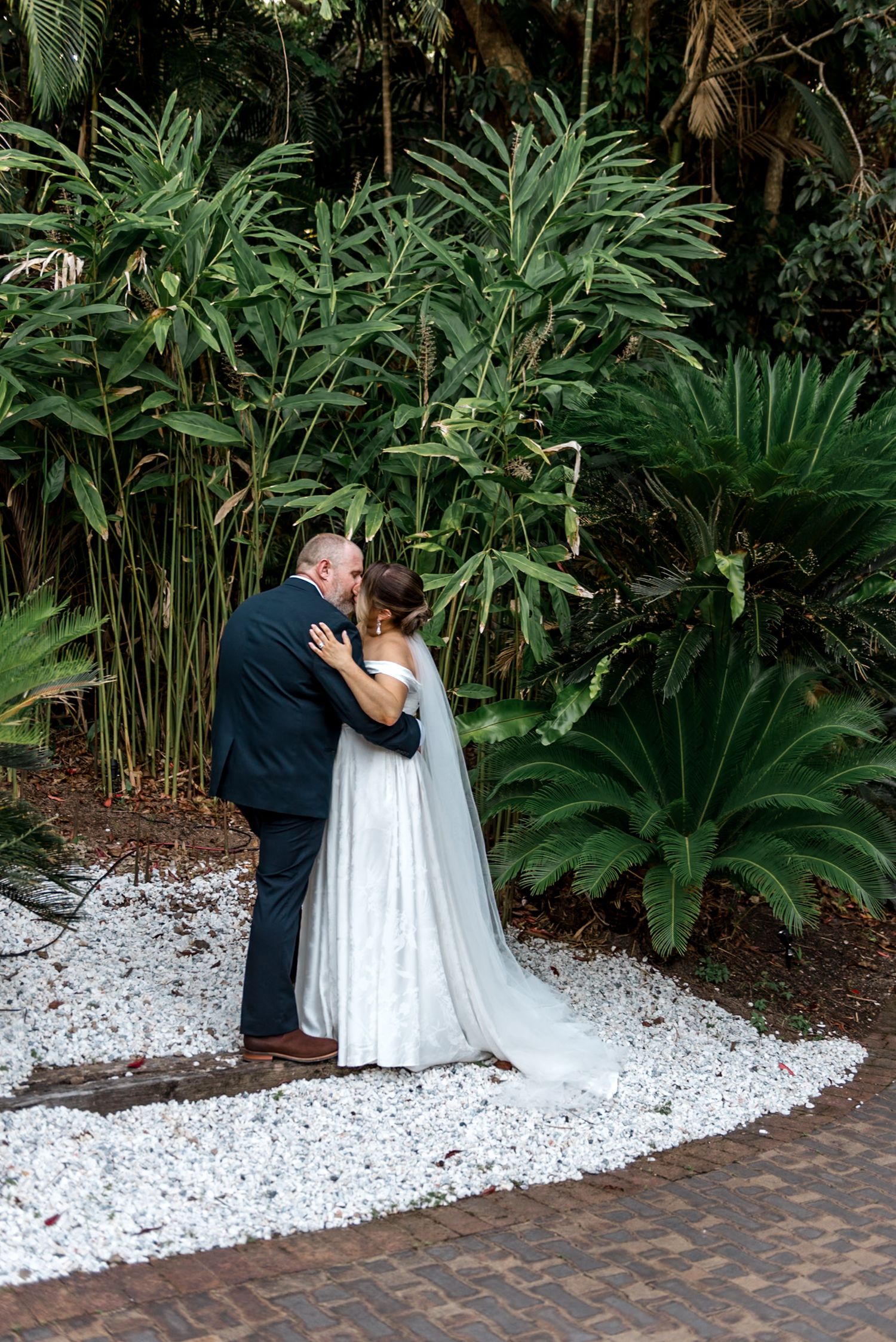Bride and groom sharing romantic kiss surrounded by lush tropical greenery on white gravel.