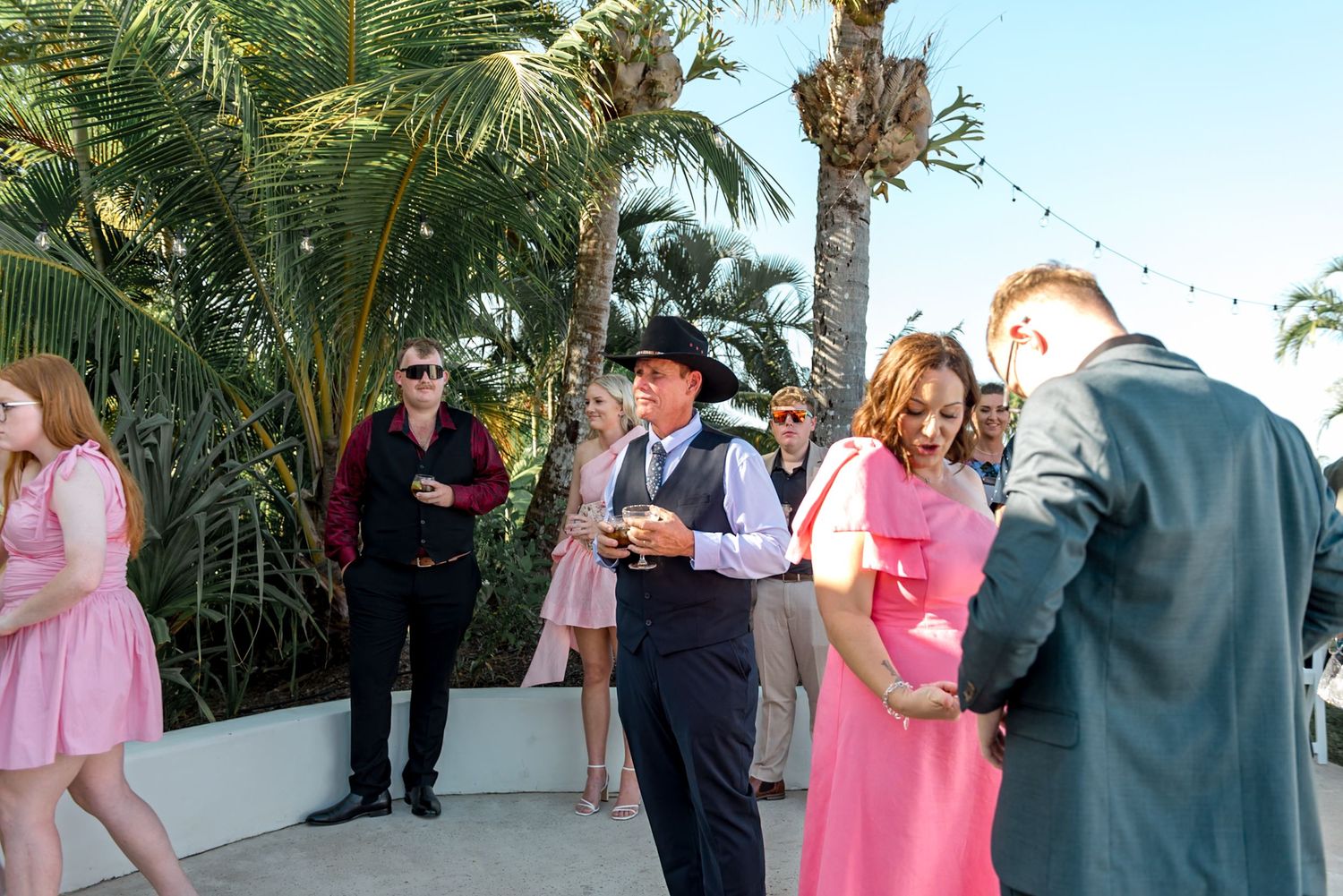 Wedding guests mingle outdoors at evening reception with string lights and tropical palms.
