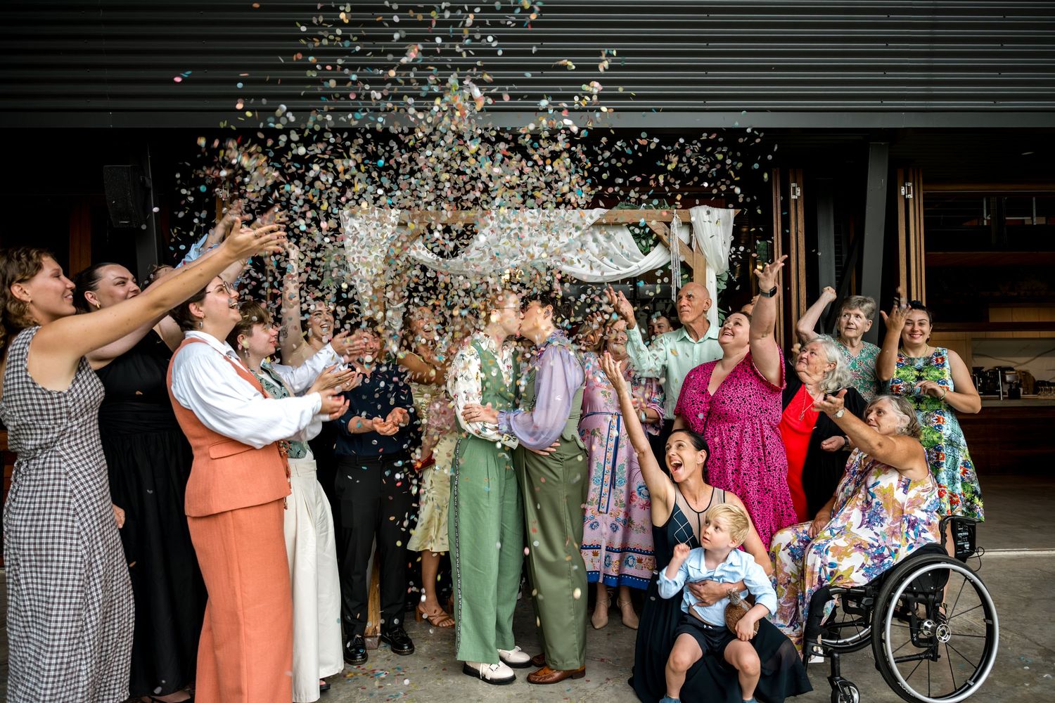 Joyful wedding couple kissing under confetti shower surrounded by cheering guests outdoors.