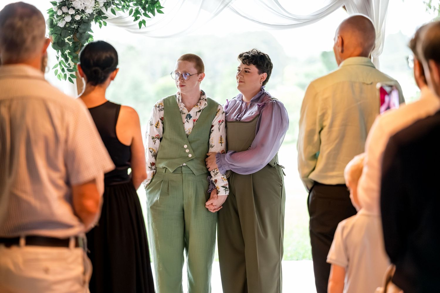 Two brides in matching green suits holding hands during outdoor wedding ceremony with guests watching.