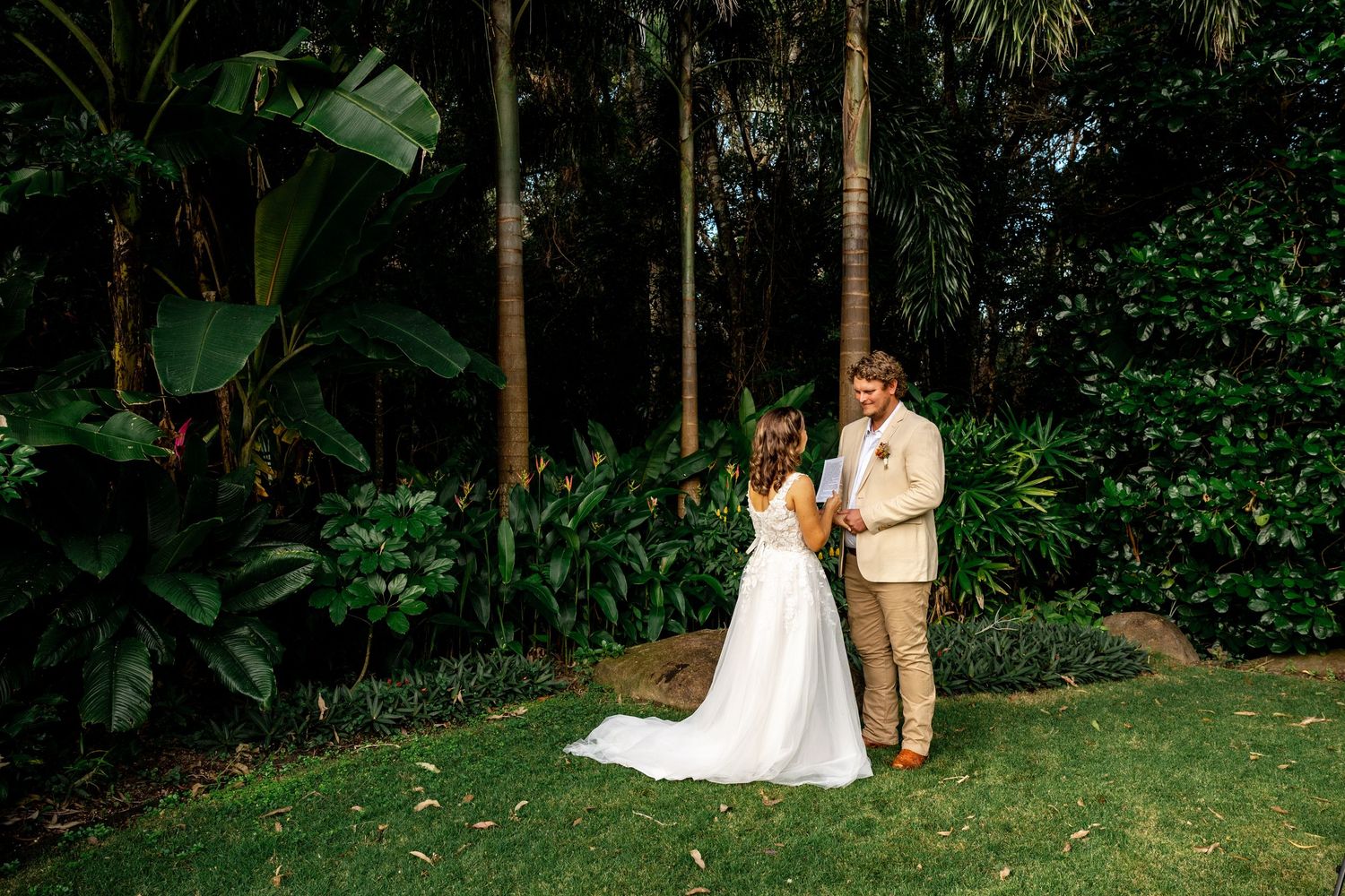 A bride and groom stand together in a lush tropical garden during their outdoor wedding ceremony.