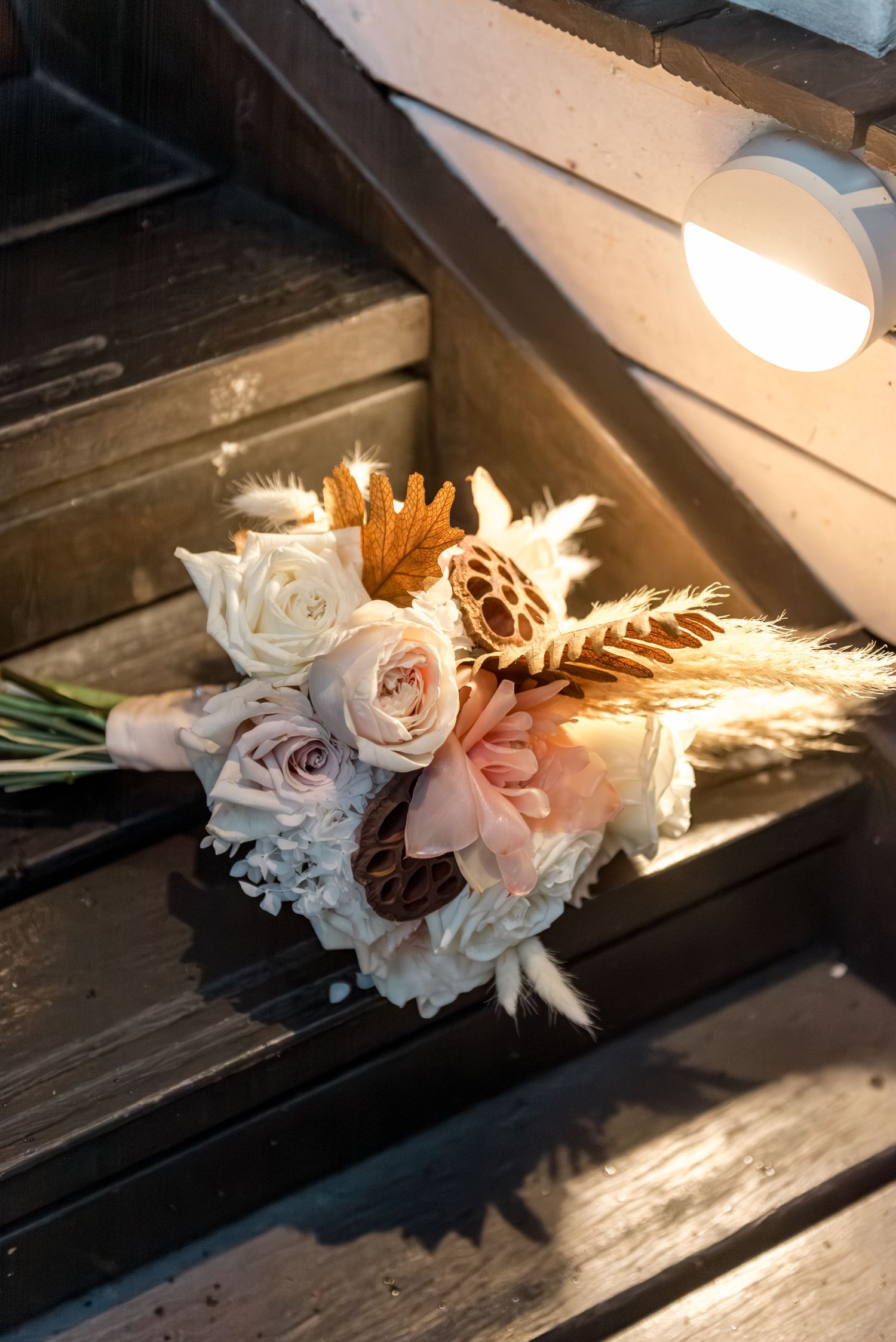 Rustic boho bridal bouquet with white roses, blush blooms, dried pampas grass, and wheat on wooden steps.