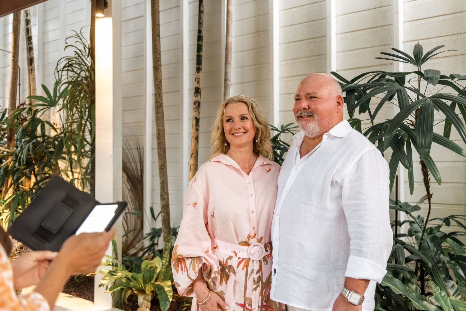 Happy mature couple smiling during an outdoor ceremony while an officiant reads from a tablet.