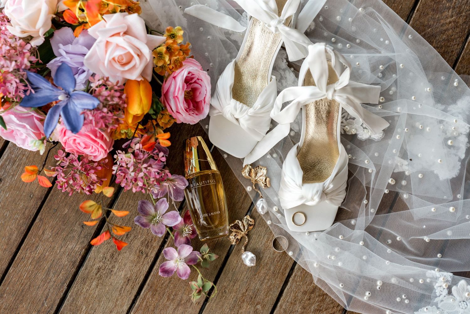 Bridal flat lay with colorful bouquet, white ribbon heels, veil, perfume bottle, and pearl earrings on wood.