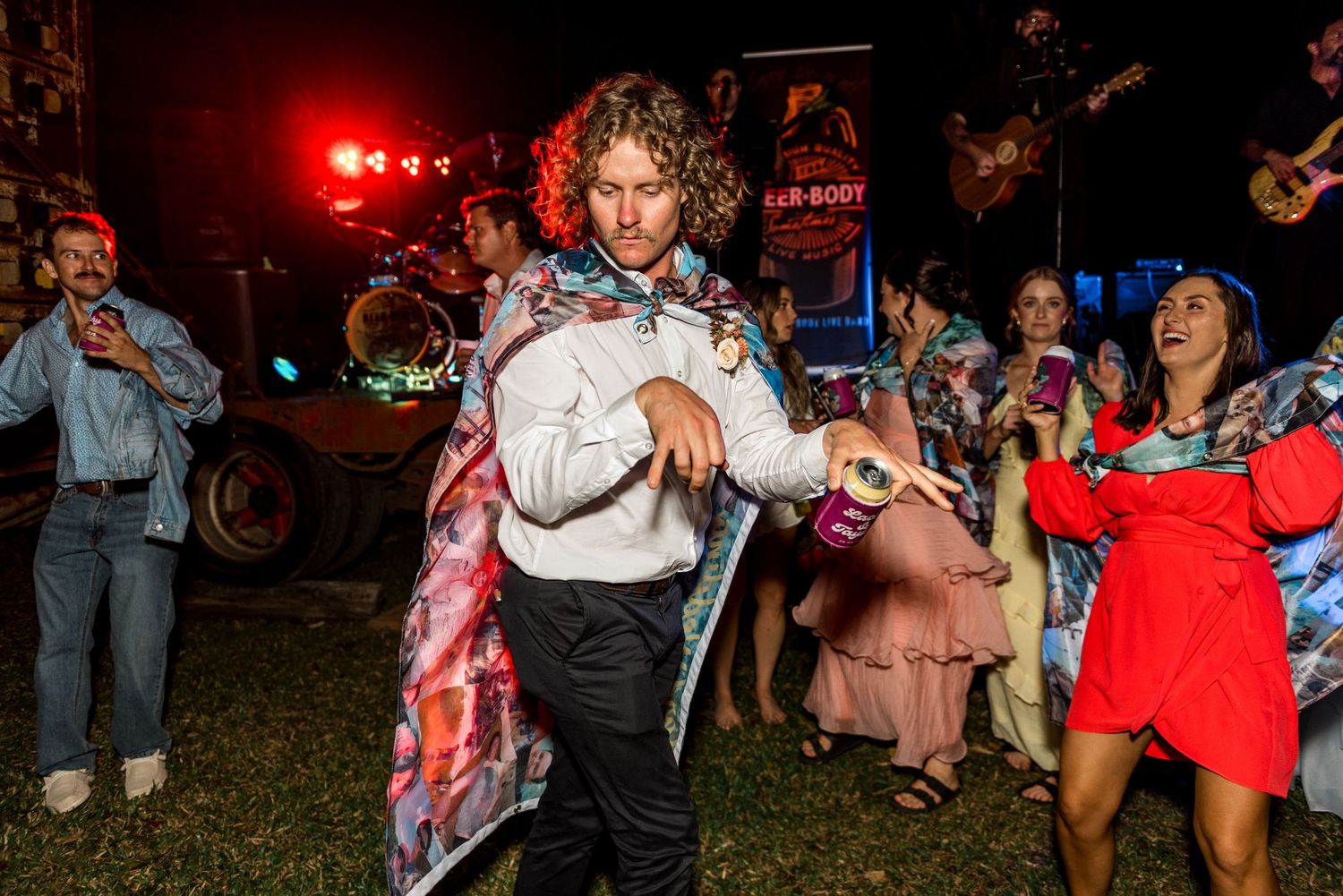 Curly-haired man in floral jacket dancing at an outdoor night wedding reception.