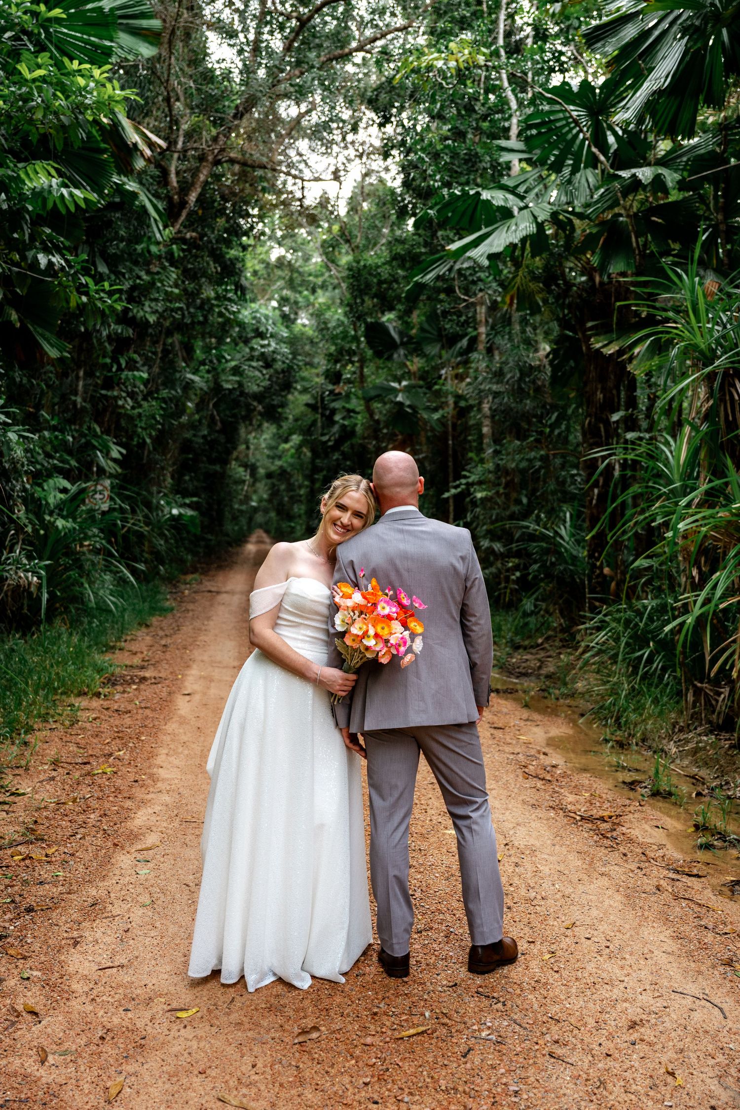 Bride leans on groom as they walk away down a lush tropical dirt road, bride holding colorful bouquet.