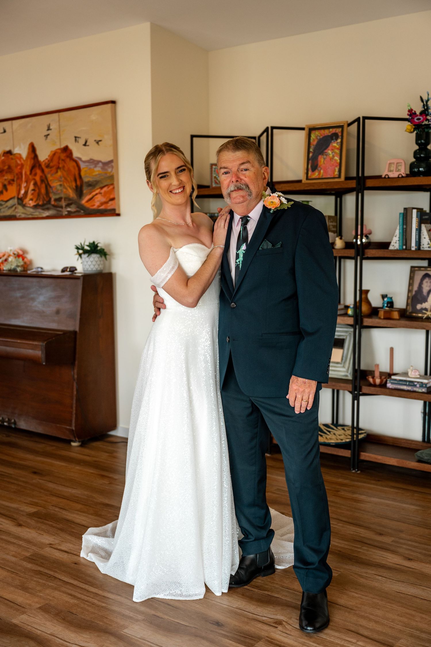 Smiling bride in white off-shoulder gown posing with father in navy suit before wedding.