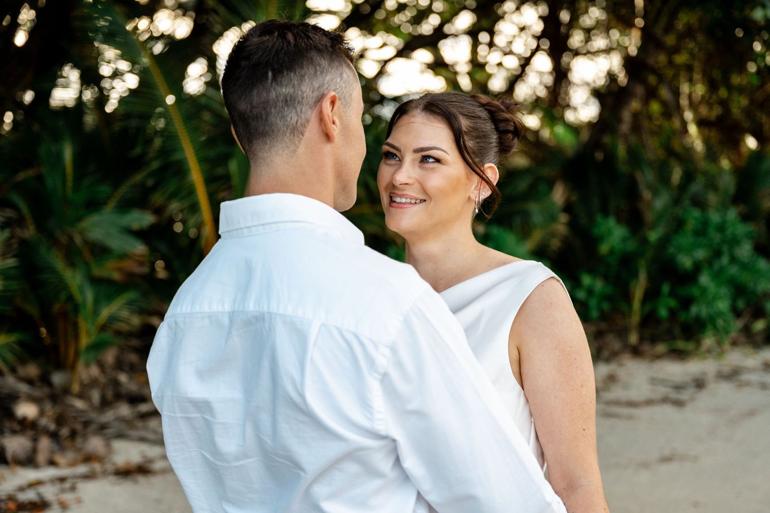 Bride in white dress smiles at groom on tropical beach with lush green foliage background at Four Mile Beach, Port Douglas.
