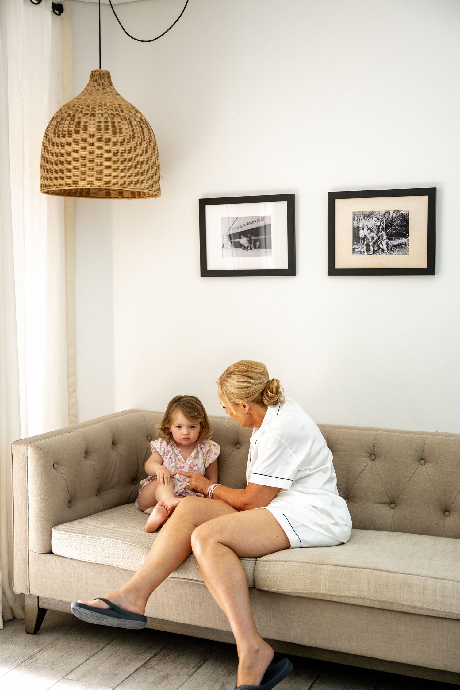 Blonde woman in white outfit sitting on a sofa helping a young toddler girl with her accessories.