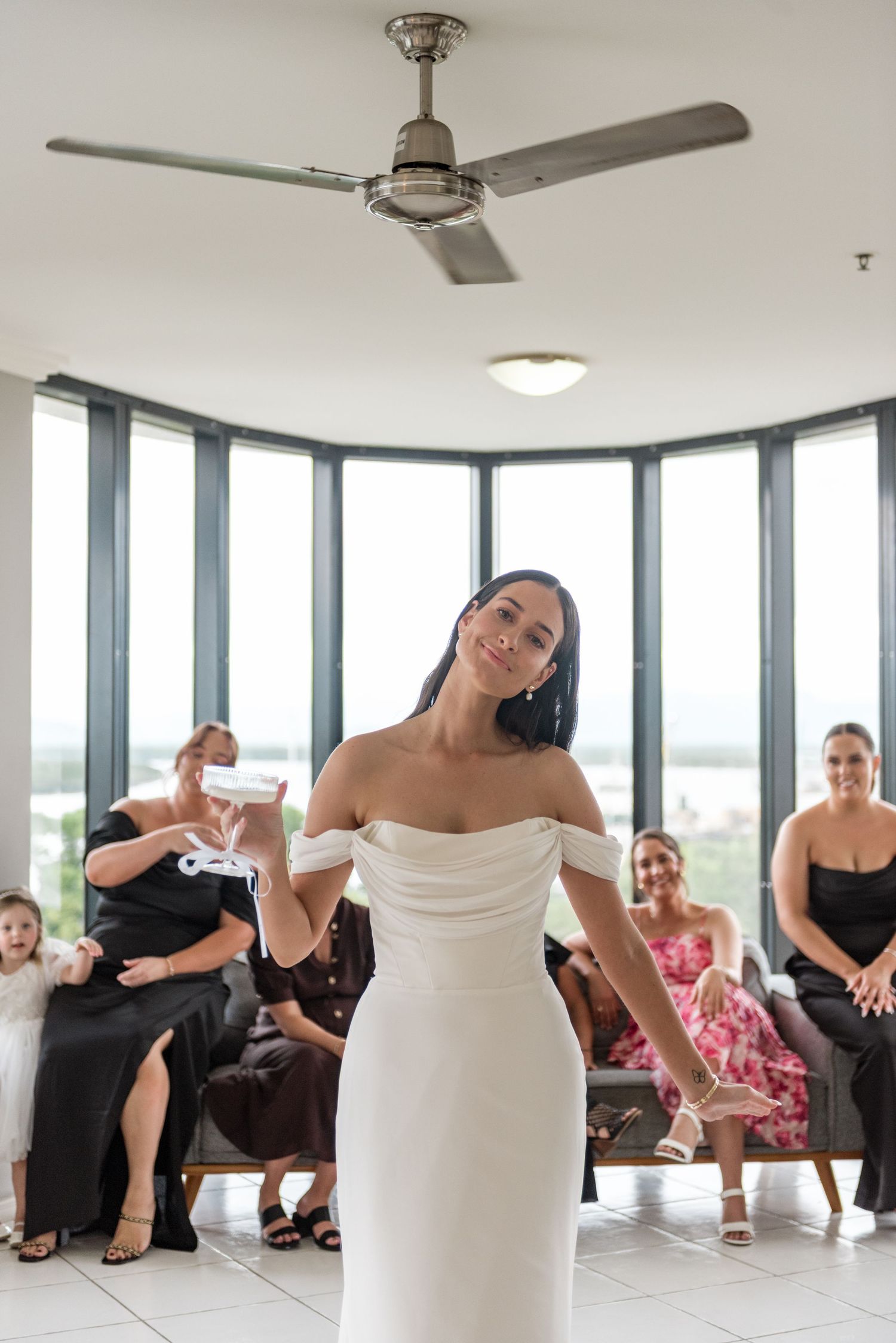 A radiant bride in a white off-shoulder fitted gown smiles and poses while guests react with joy behind her.