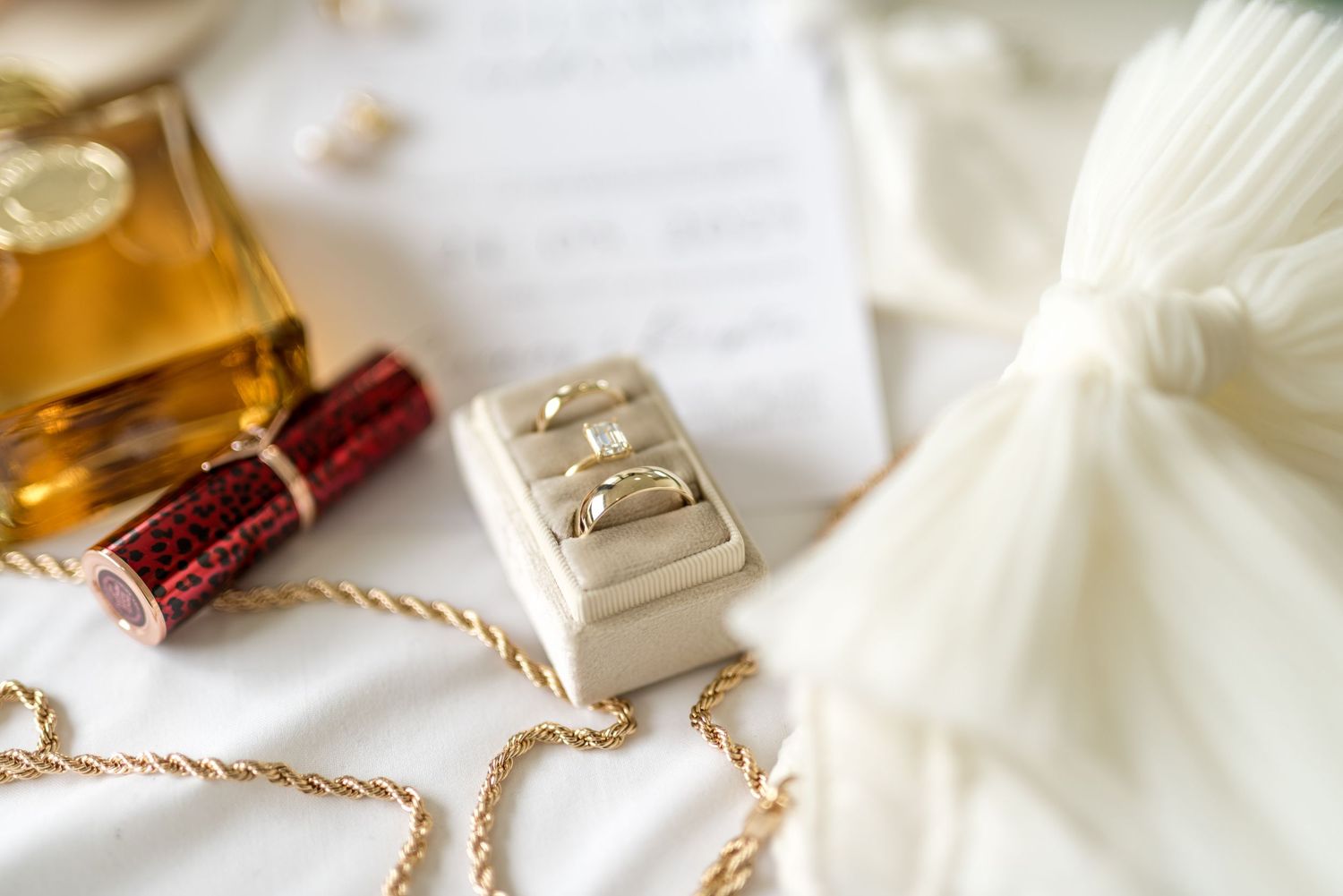Close-up of gold wedding rings in a velvet box surrounded by perfume, lipstick, and pearl chain on white fabric.