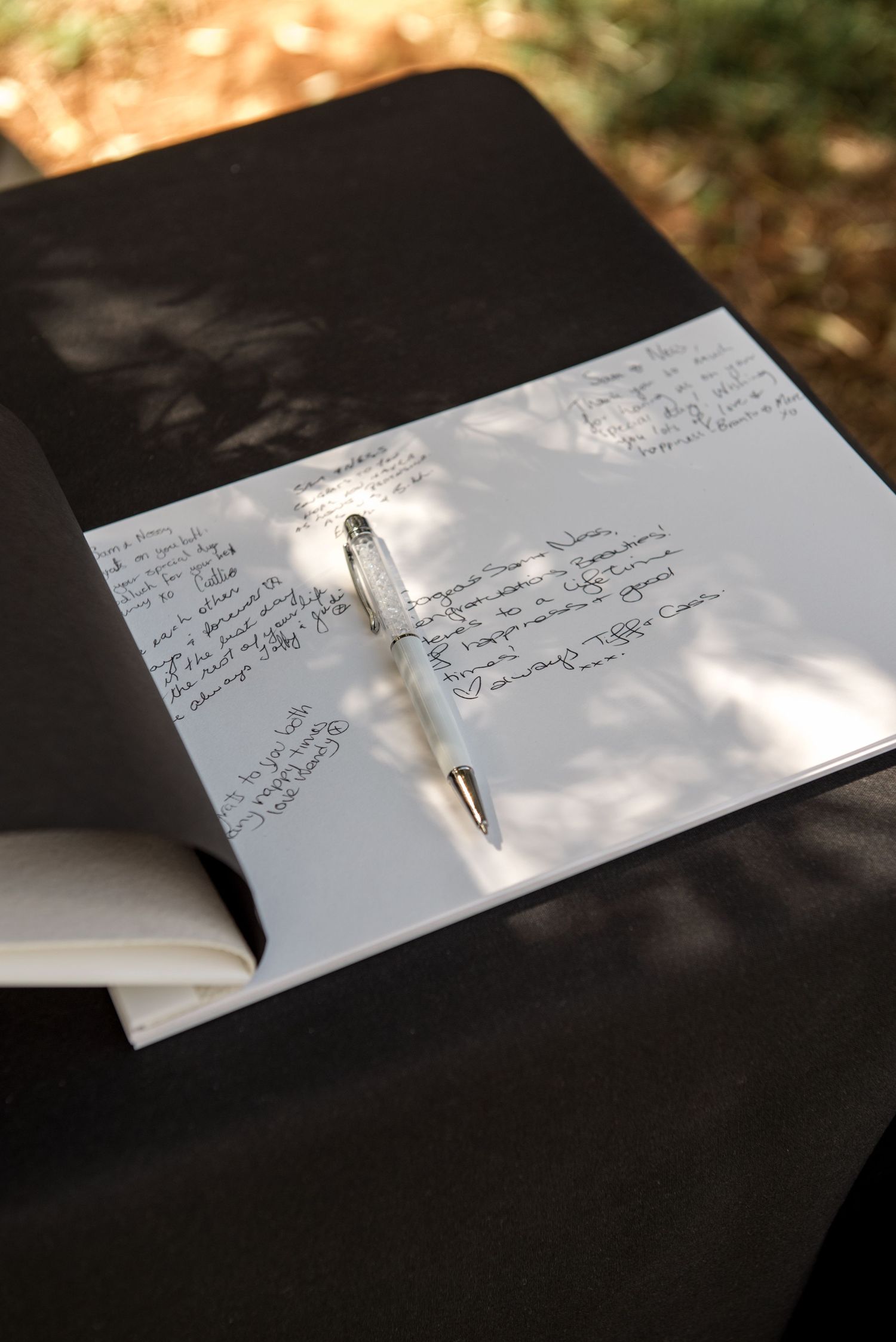 Open wedding guest book with handwritten messages and a pearl pen on a black draped table outdoors.