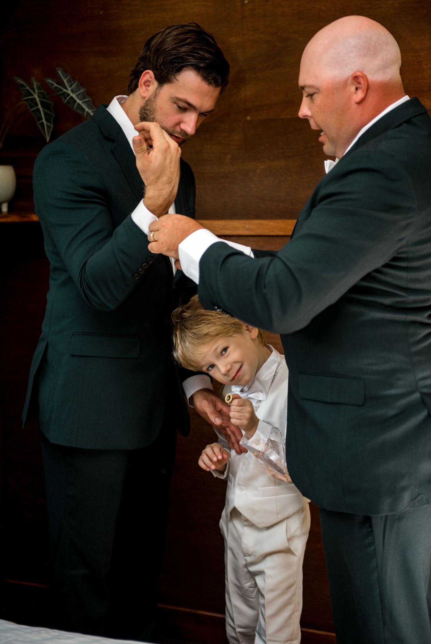 Two groomsmen in dark suits help young ring bearer with his outfit in a wood-paneled room.