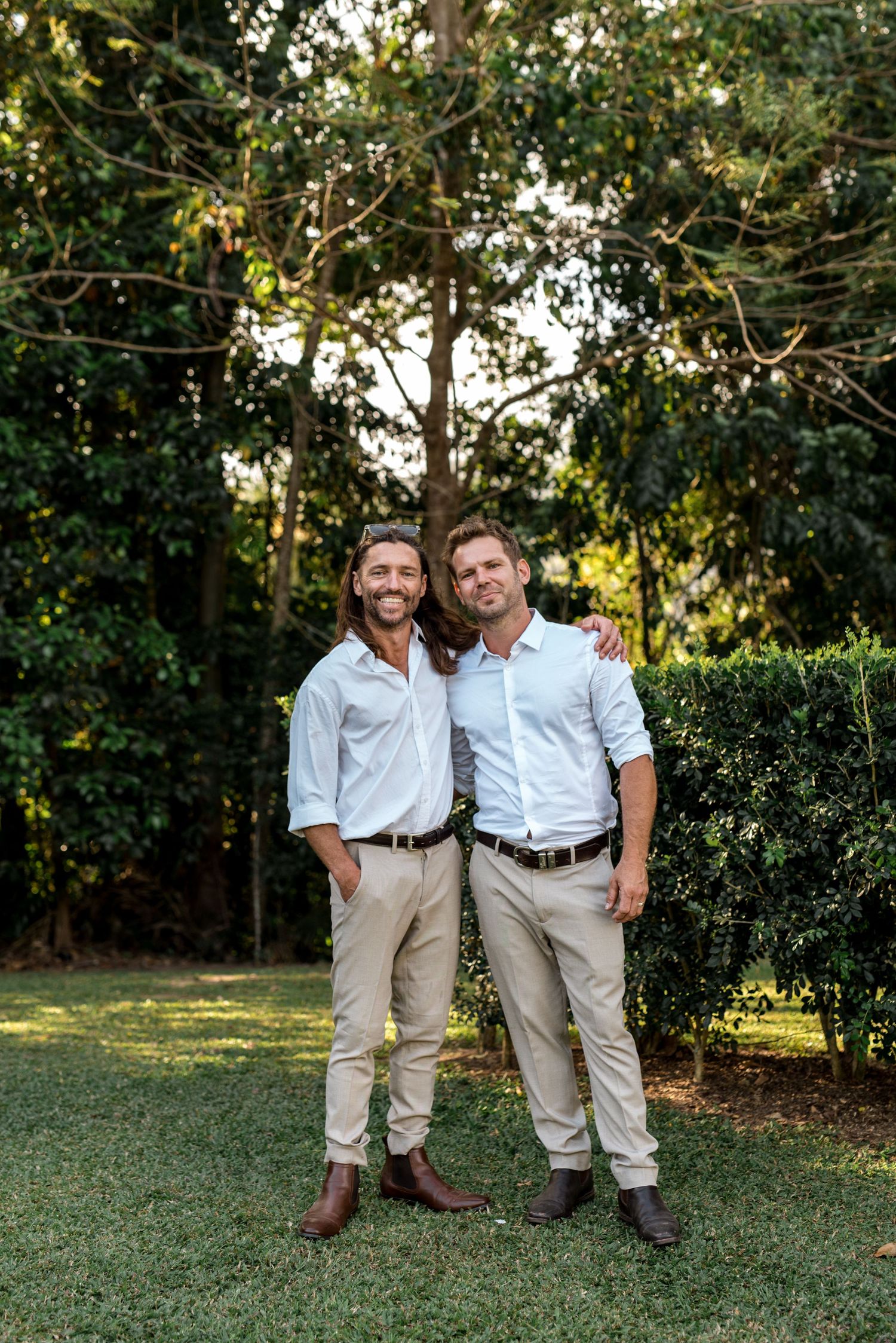 Two groomsmen in white shirts and tan trousers smile together in lush garden wedding setting.