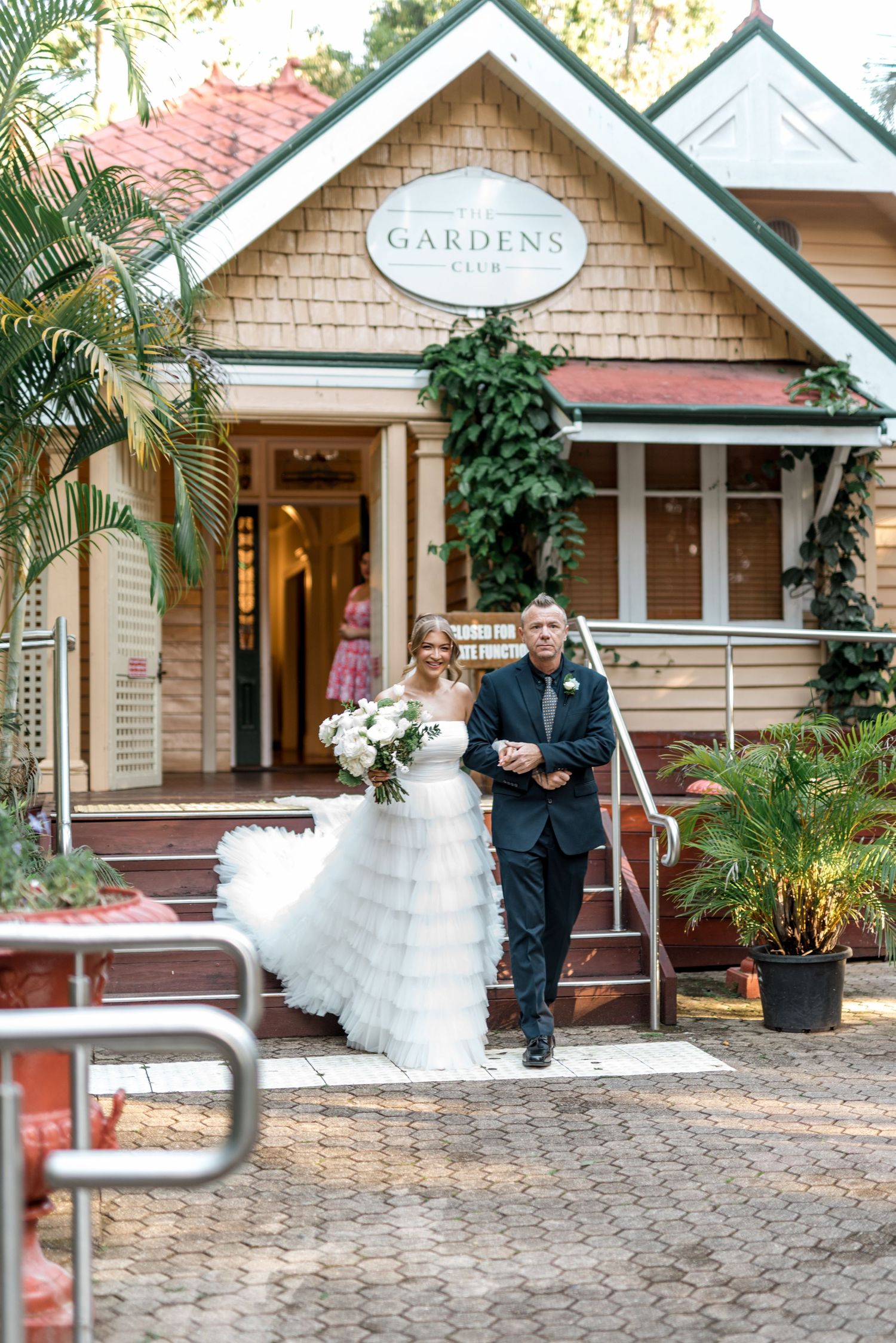 Bride in layered tulle gown walks with escort from Gardens Club venue entrance.
