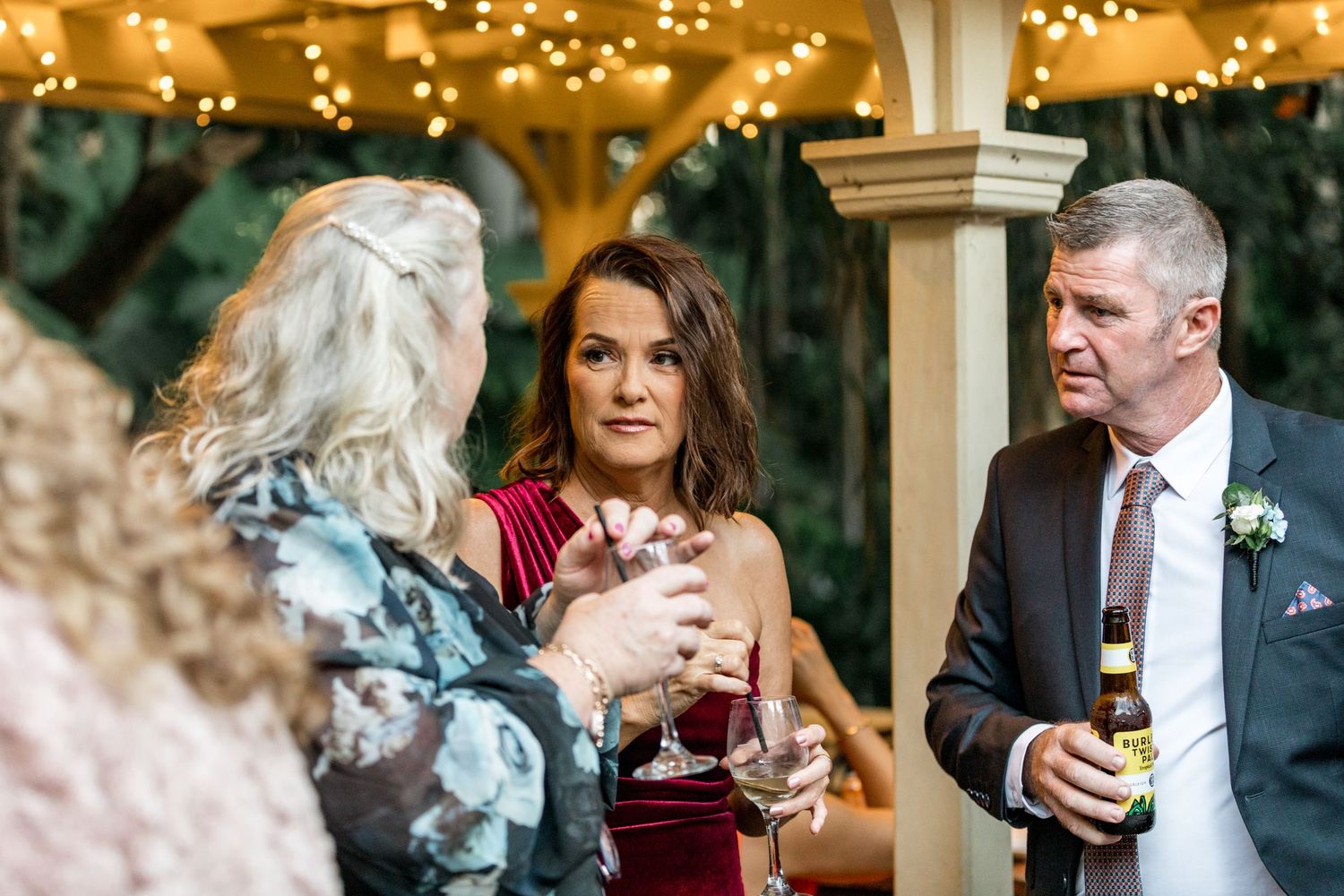 Elegantly dressed wedding guests mingle and clink glasses under a pergola adorned with warm fairy lights.