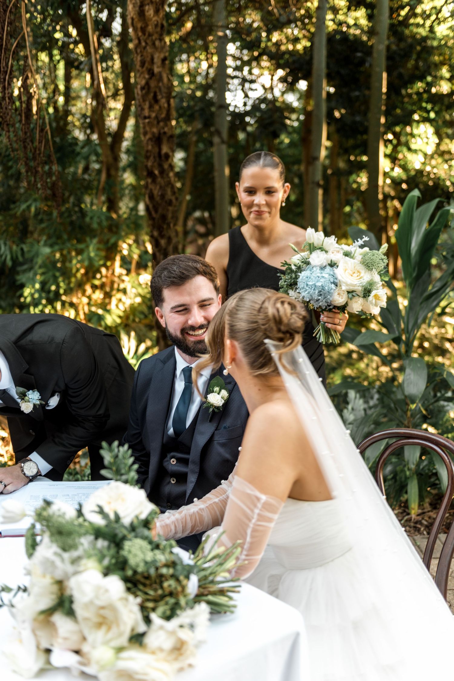 Laughing groom sits at signing table with smiling bride and bridesmaid in black at garden wedding.