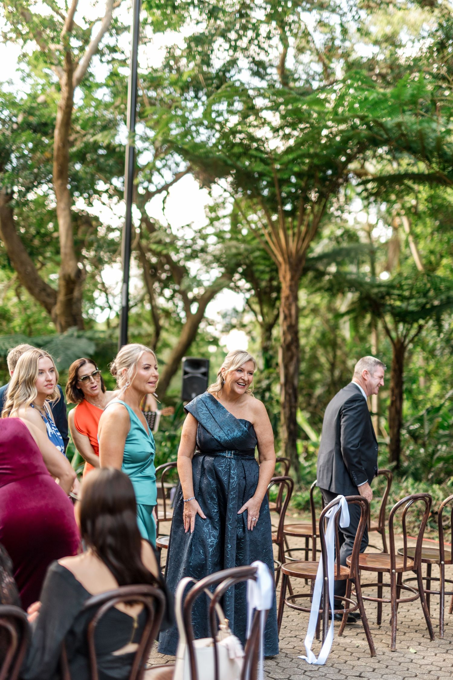 Guests arrive at an elegant outdoor wedding ceremony surrounded by lush green trees.