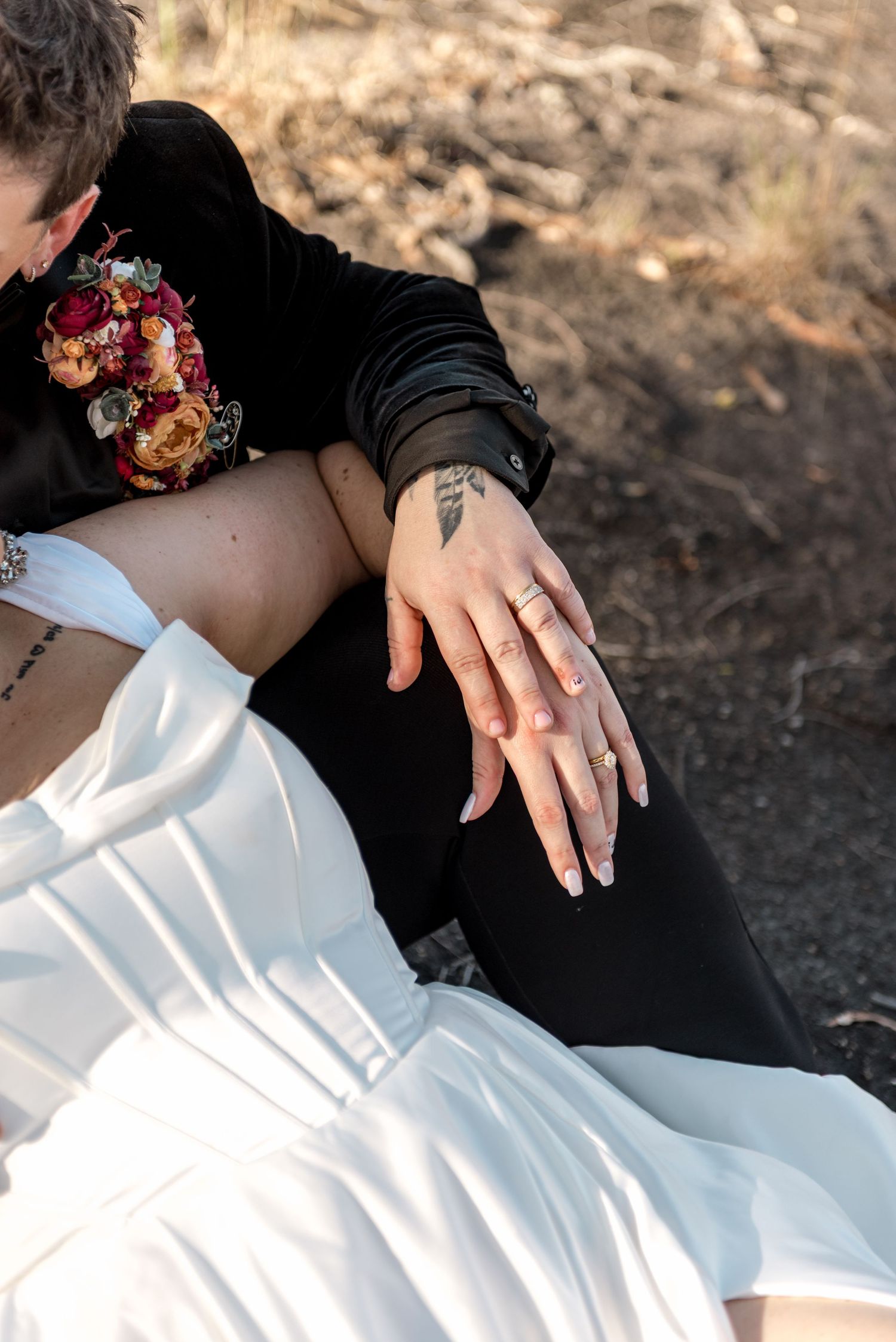 Close-up of same-sex couple's hands showing wedding rings and tattoos, with floral bouquet detail on black velvet jacket.