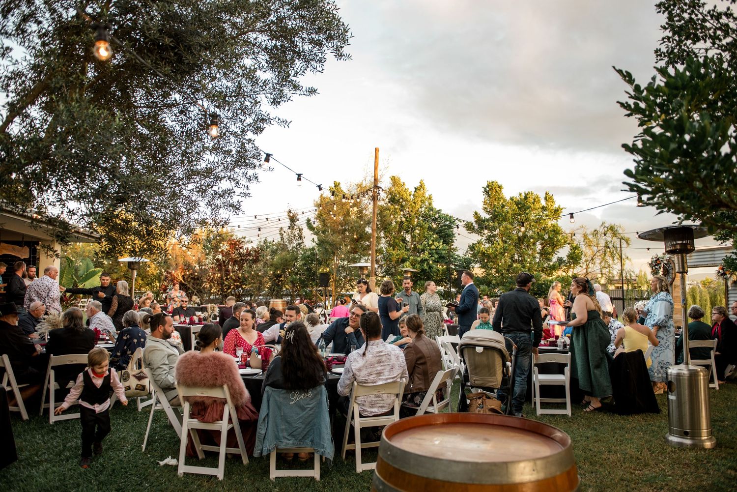 Outdoor wedding reception with guests seated at decorated tables under string lights at dusk.
