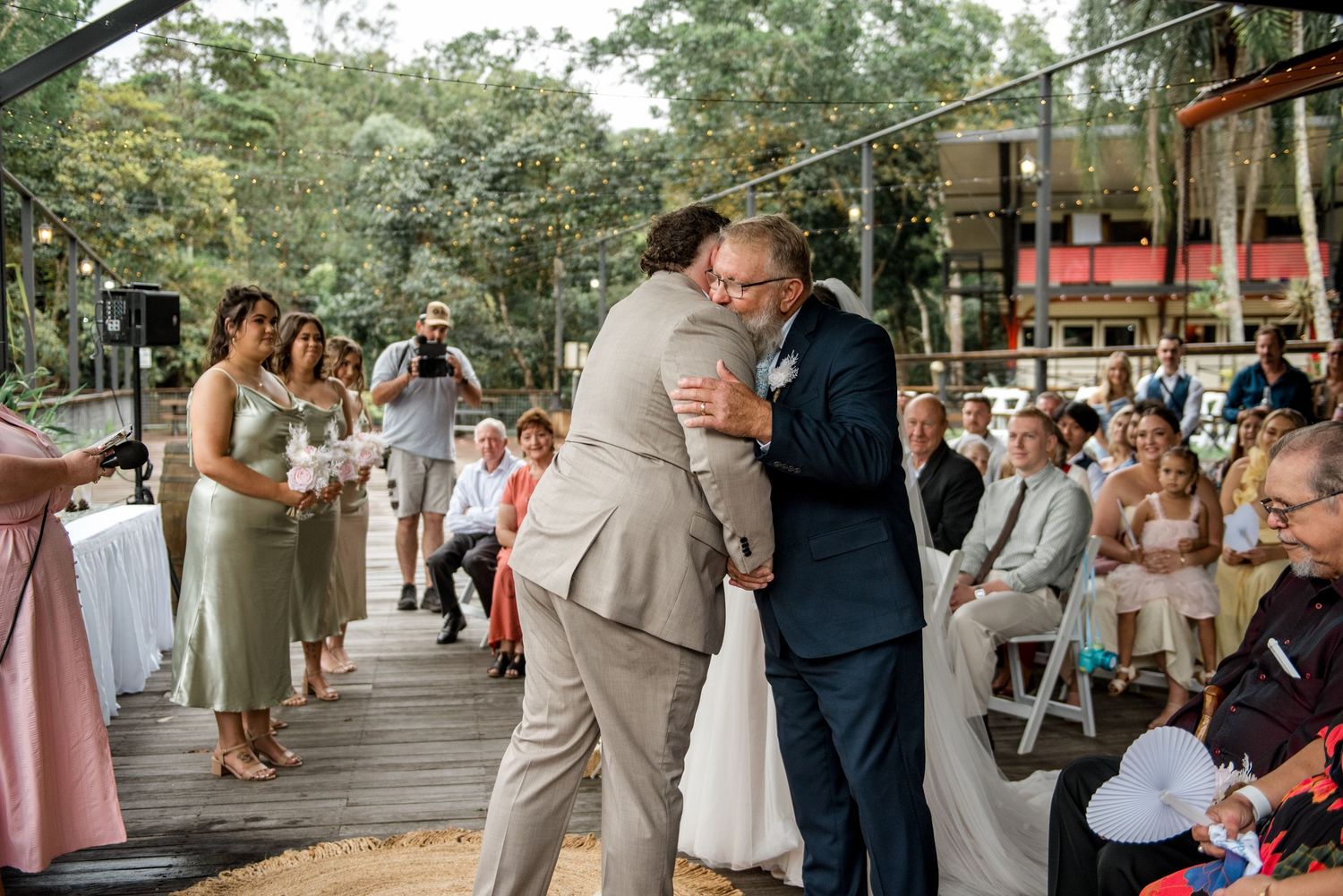 Groom in tan suit shares emotional hug with man in navy suit during wedding ceremony.