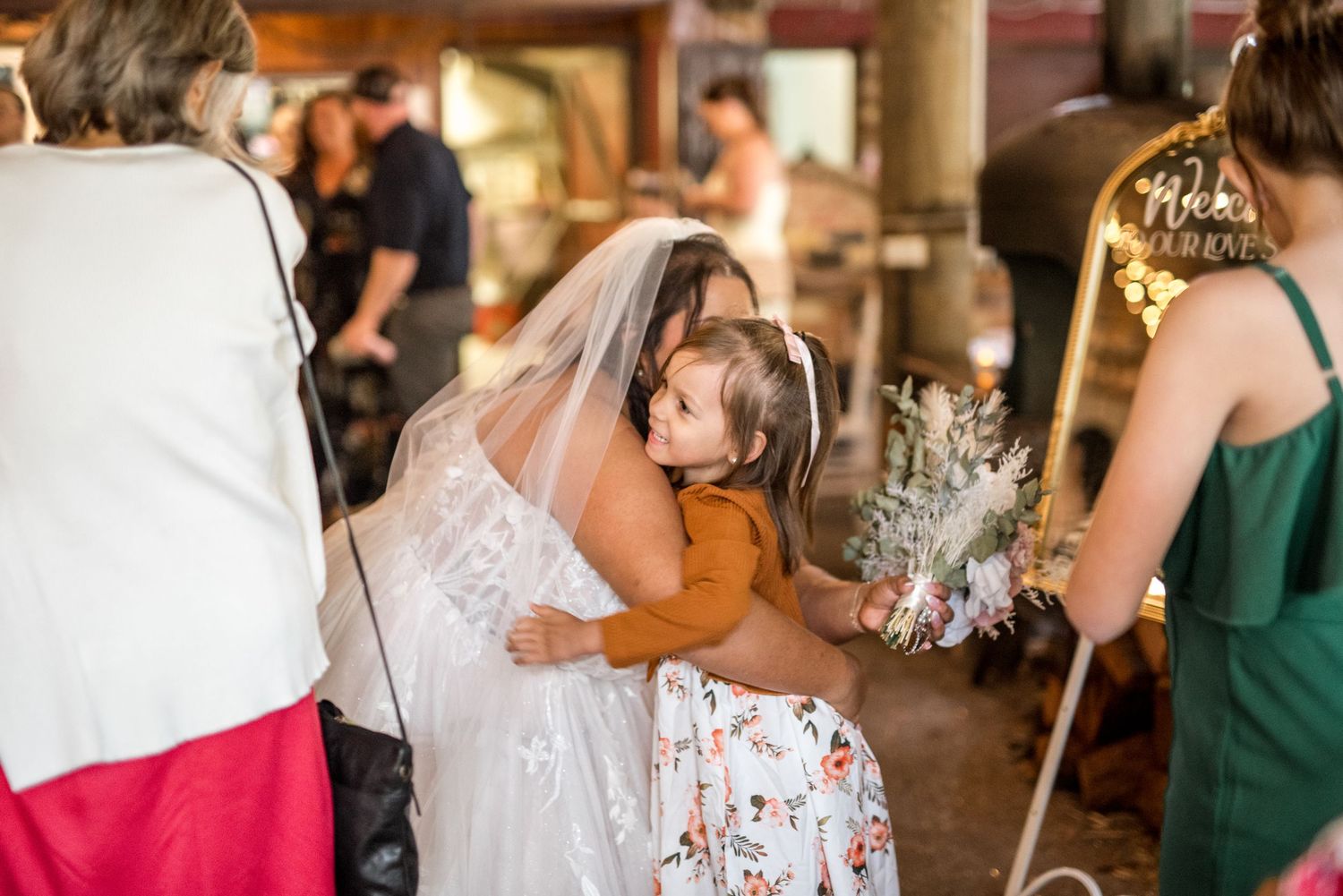 Bride in white veil bends down to hug a young child in a floral dress at wedding reception.
