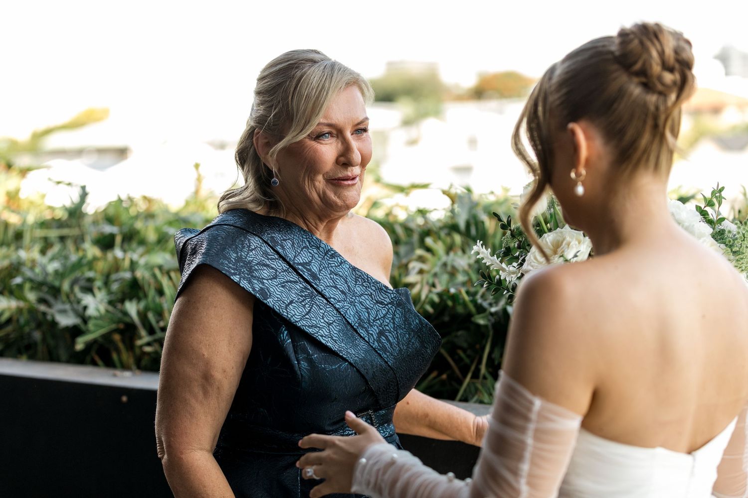 Older woman in navy formal gown sharing warm smile with bride in off-shoulder wedding dress.