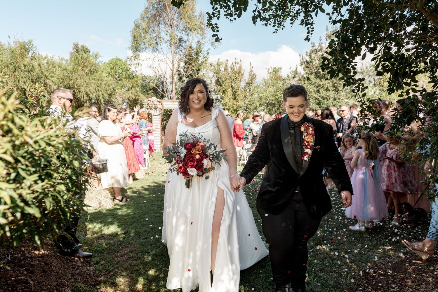 Newlywed couple walks joyfully through confetti shower surrounded by cheering guests at garden wedding.
