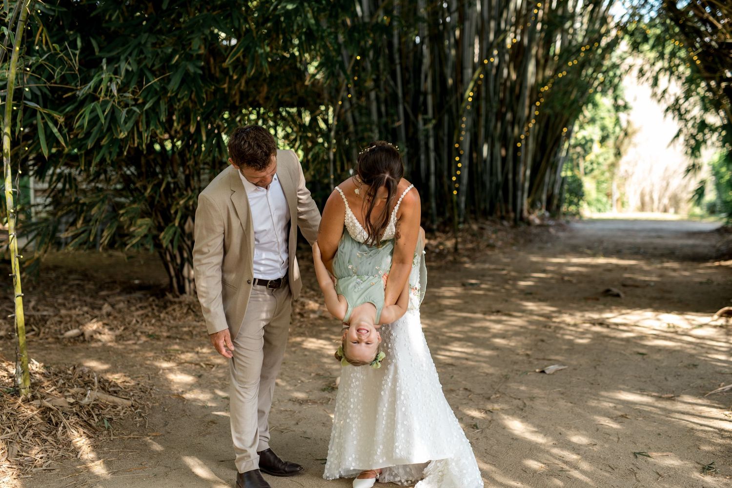 Wedding couple playfully swinging young child between them on sandy path lined with bamboo trees.