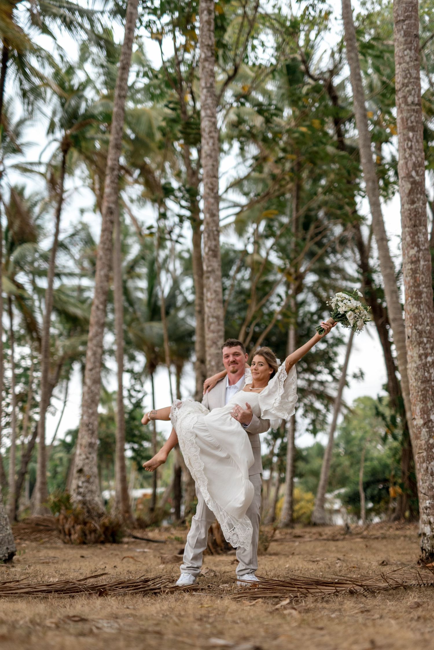Groom carrying bride in white lace gown among palm trees, bride joyfully raising bouquet in the air.