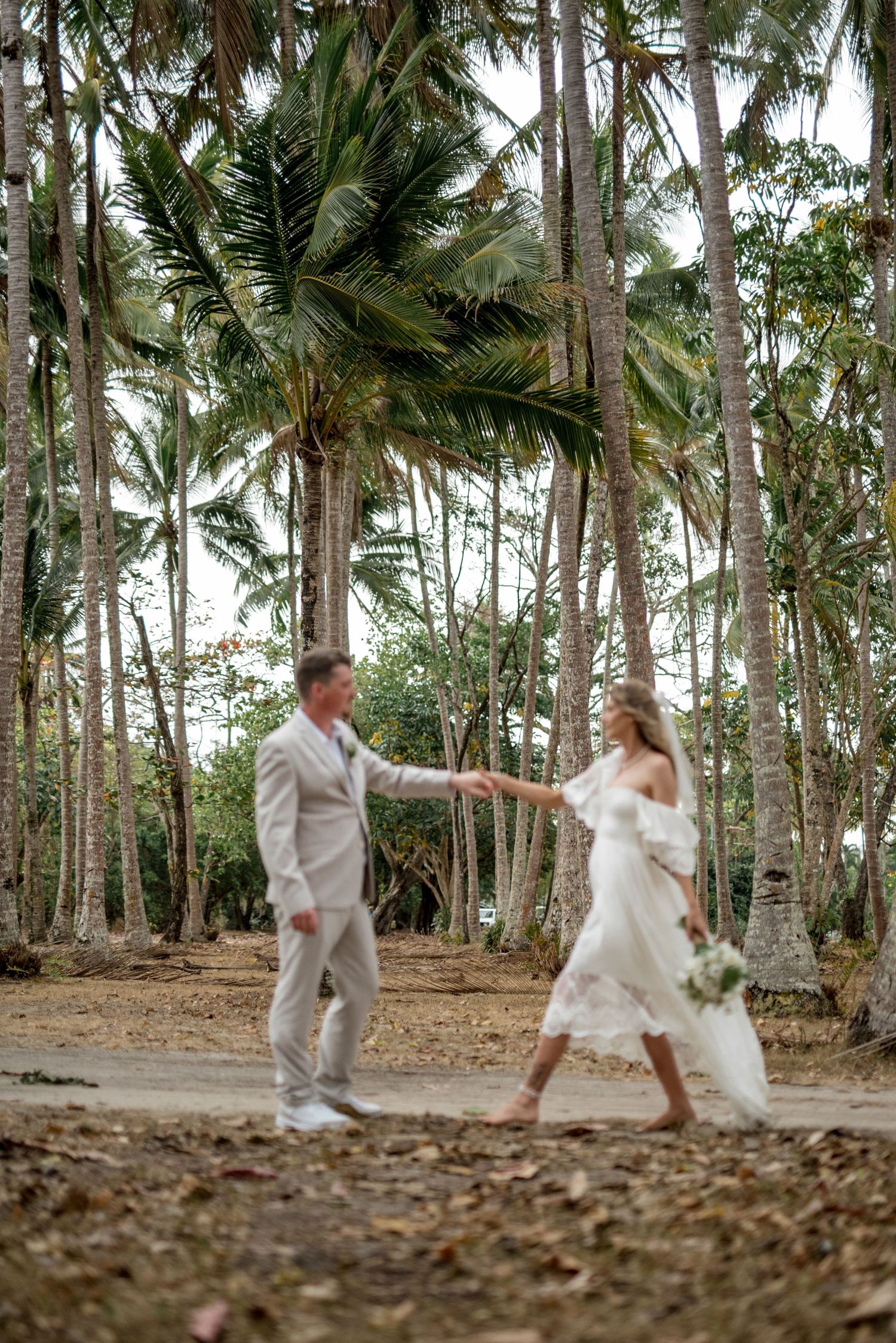Bride and groom walk hand in hand through tropical palm tree grove during wedding photo session.