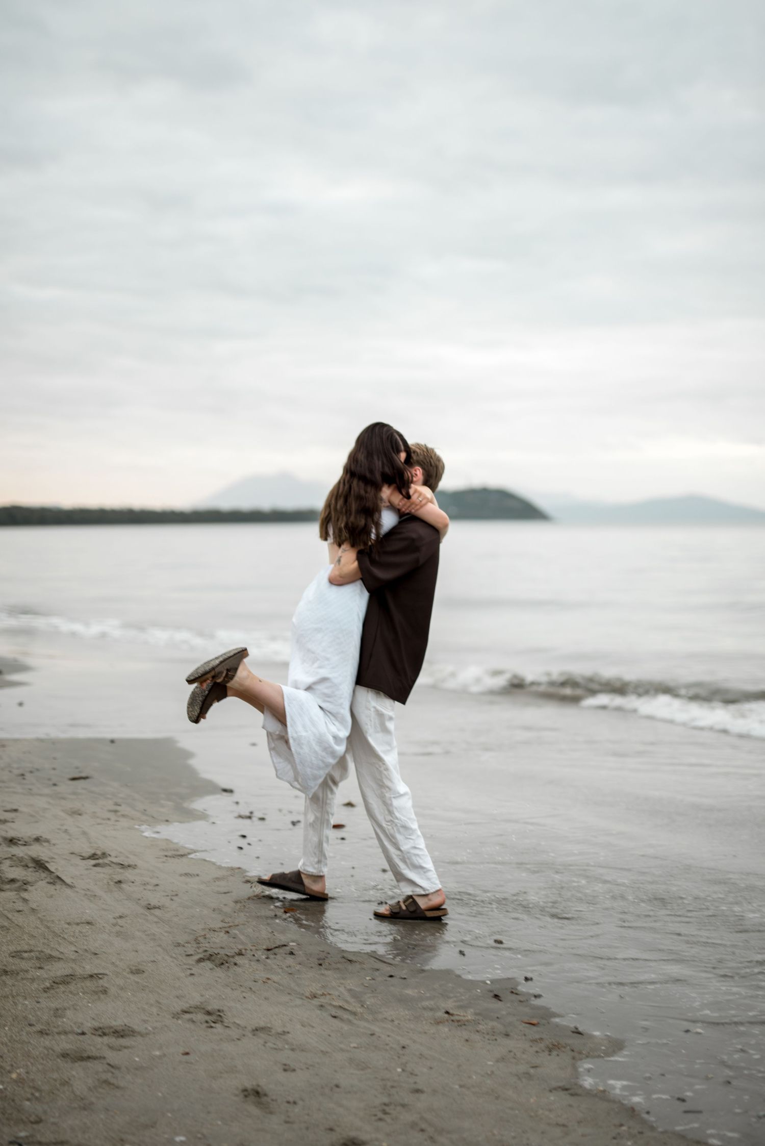 Man lifts woman in joyful embrace on wet sandy beach at dusk.
