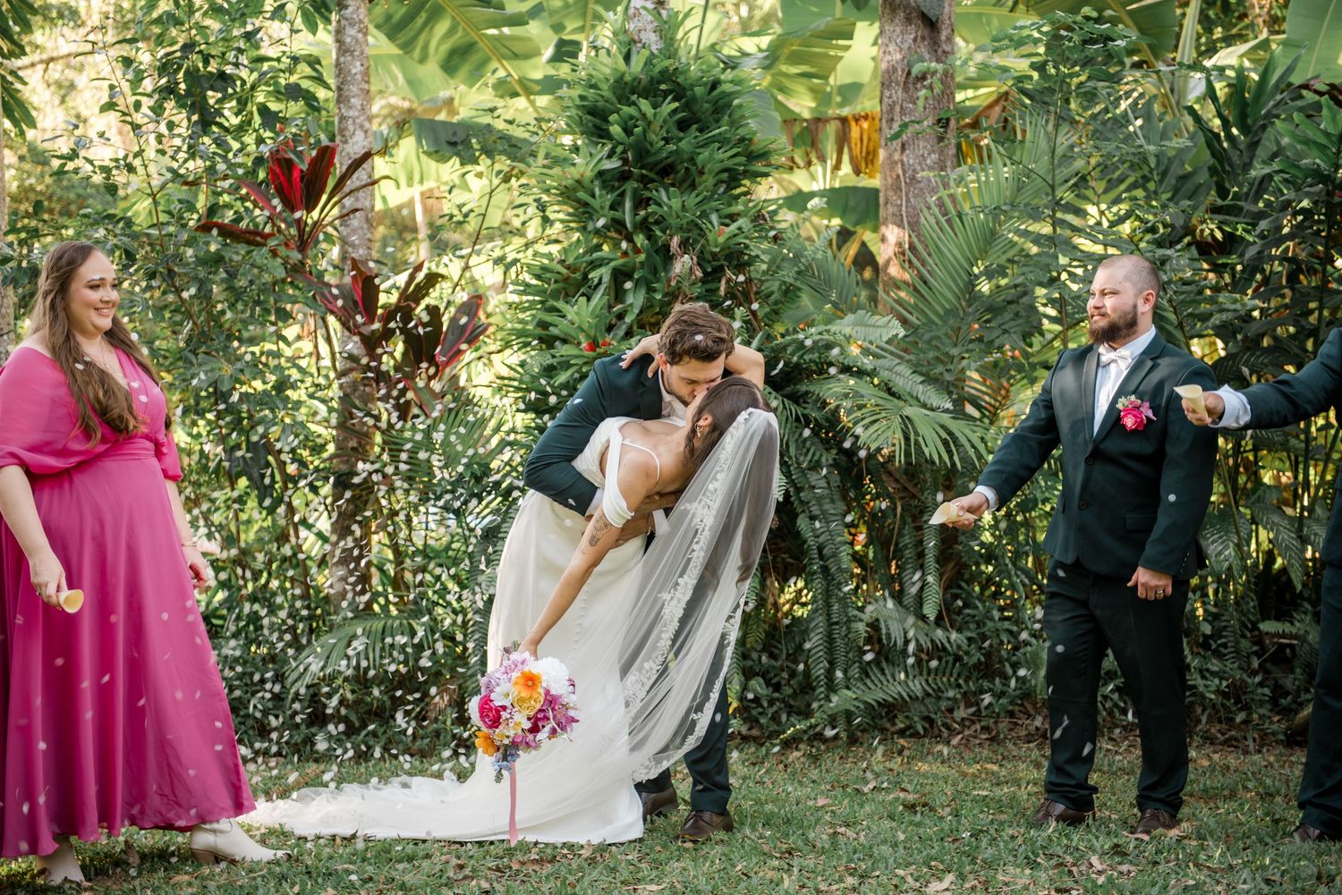 Groom dips bride for a kiss while bridesmaid in pink and groomsman watch during outdoor wedding ceremony.