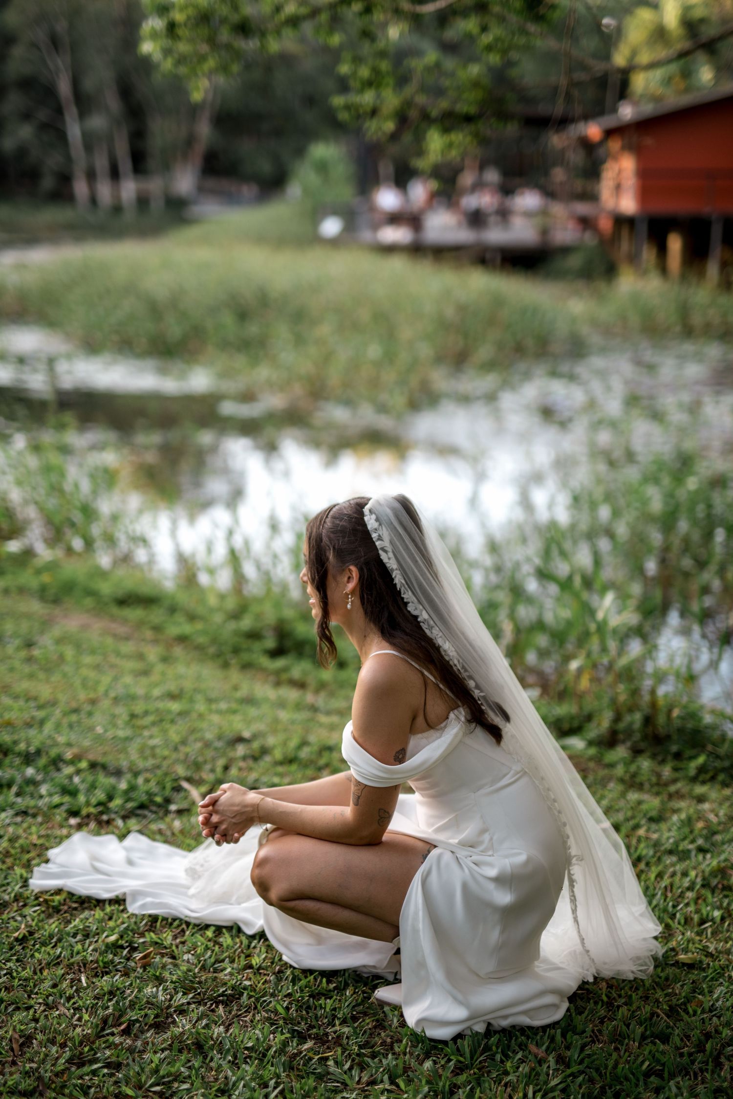 Bride in white off-shoulder gown and veil sits gracefully beside a peaceful reflective pond.