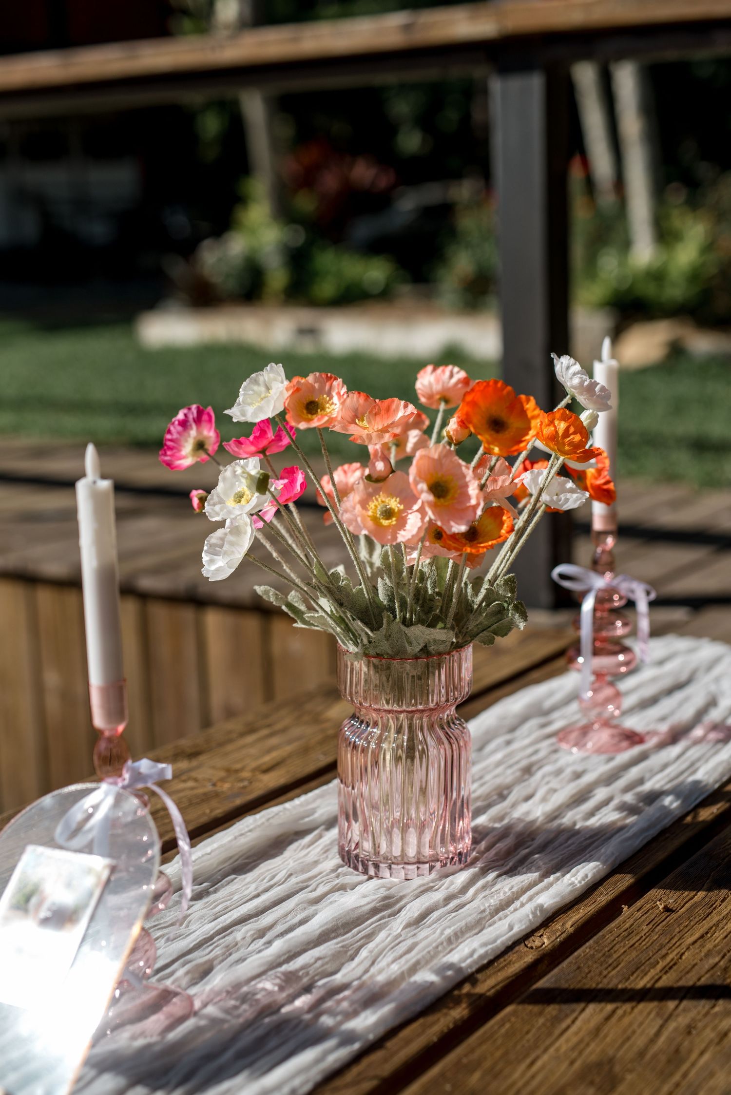 Pink ribbed glass vase filled with colorful orange and pink poppies on a rustic wooden table with candles at a wedding.