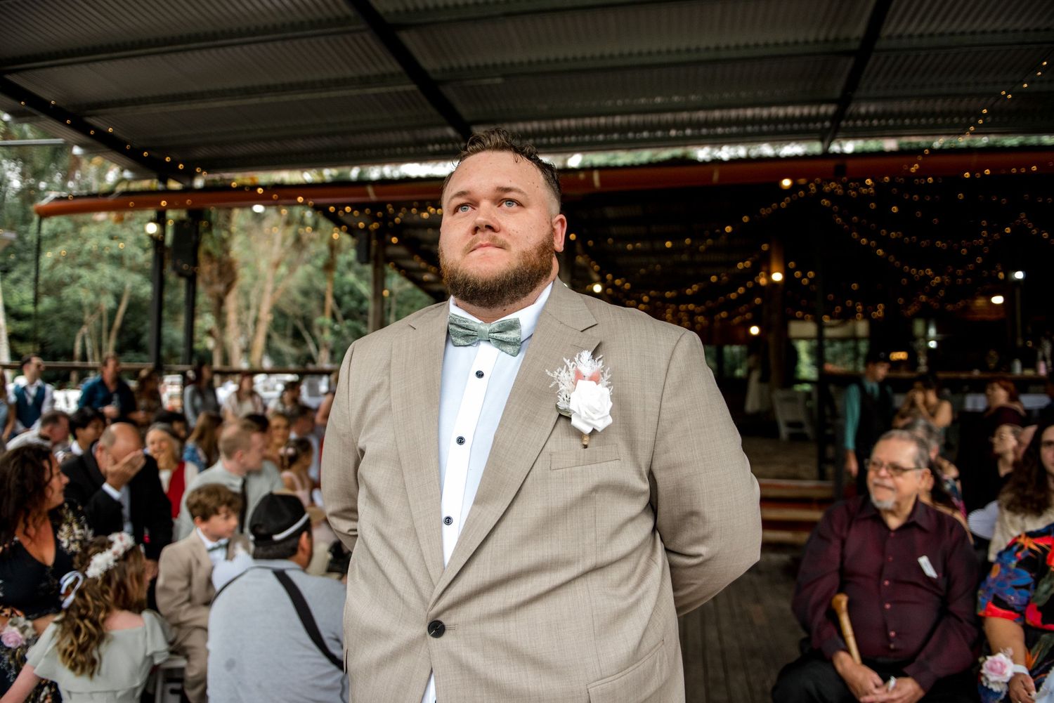 Groom in tan suit with teal bow tie and boutonniere stands at outdoor wedding ceremony.