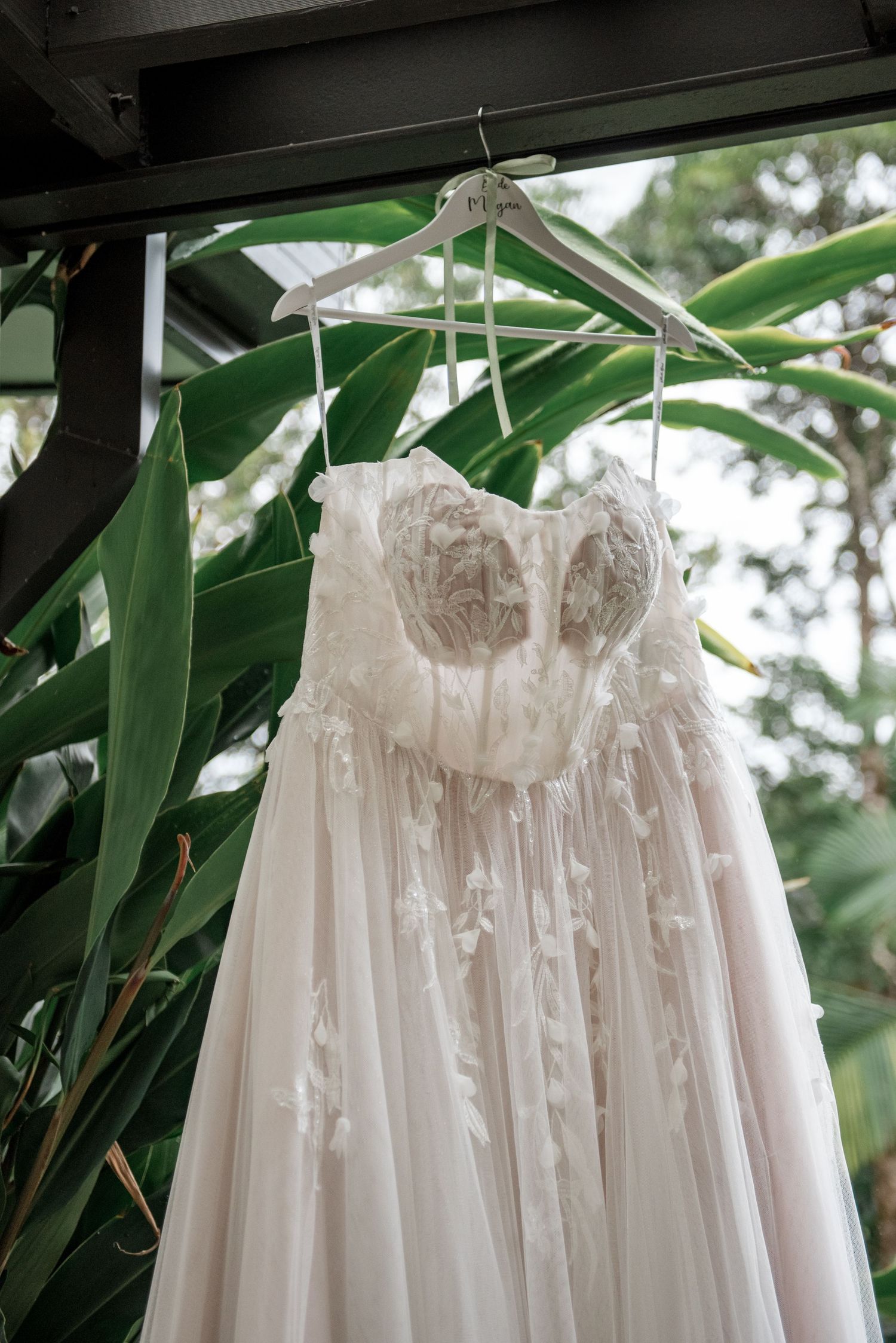 Elegant strapless lace wedding dress hanging on a personalized hanger against tropical green foliage.