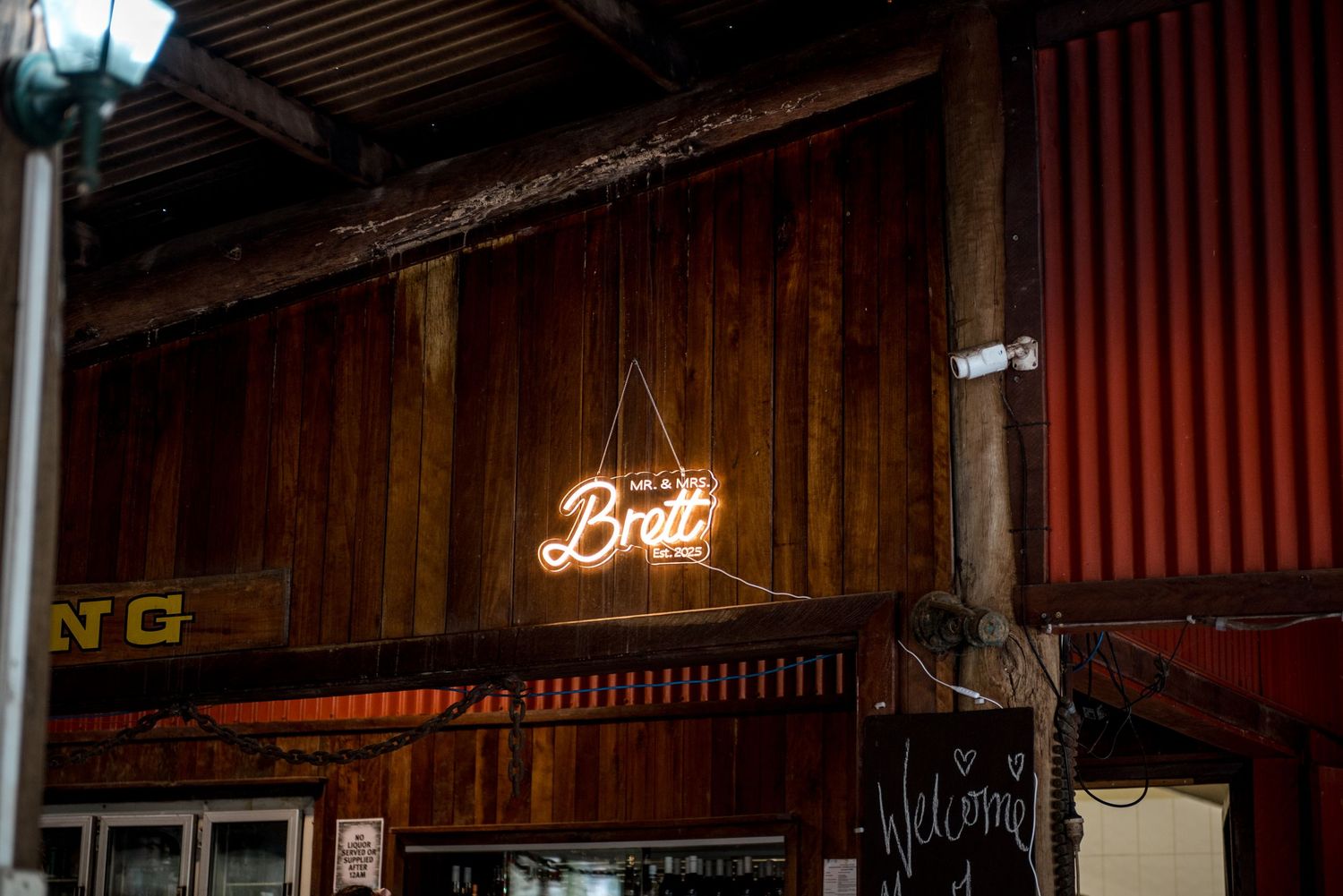 Neon sign displaying a name Brett hanging on a rustic corrugated iron wall at a wedding venue.