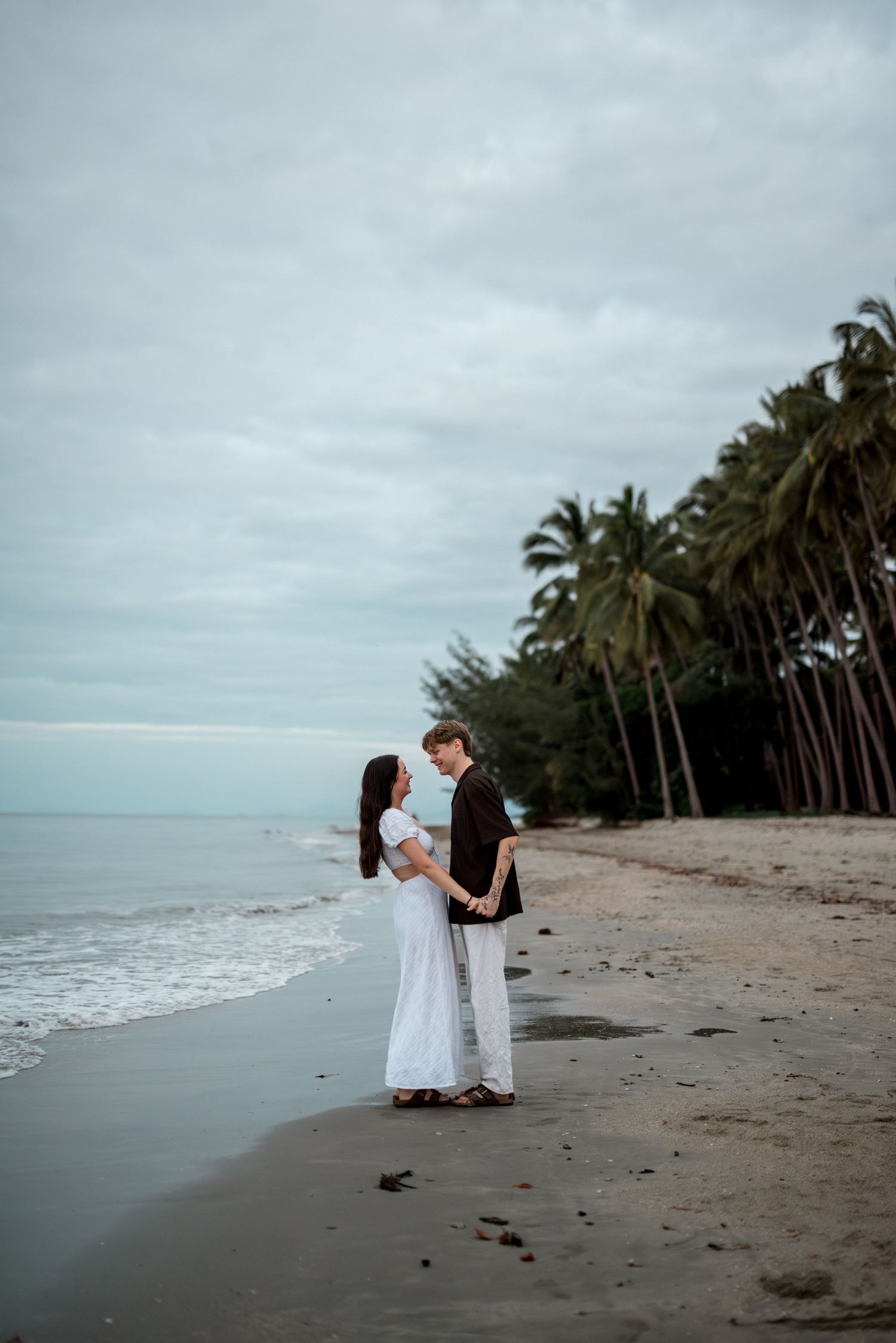 Couple holding hands facing each other on a tropical beach with palm trees and overcast sky.