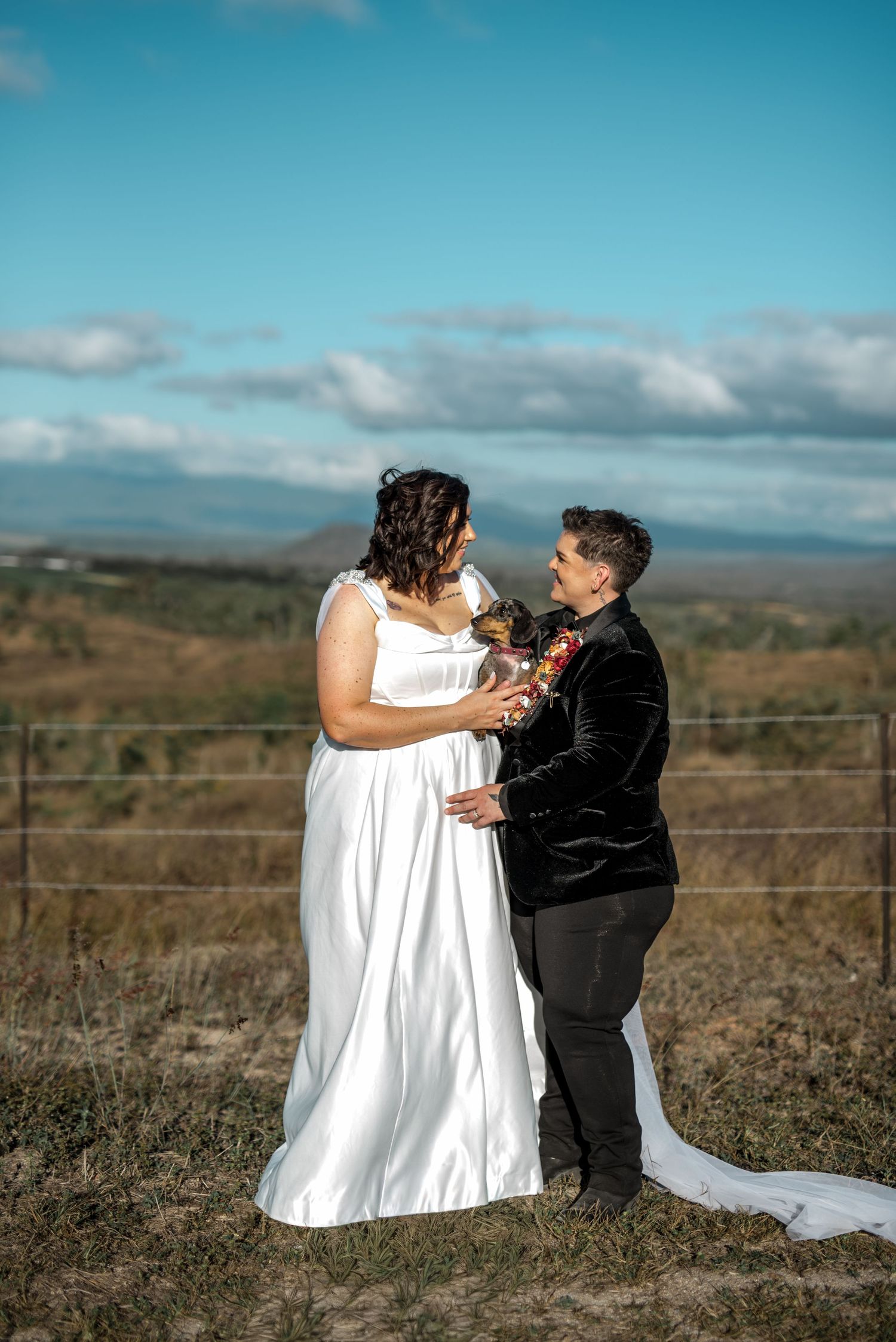 Same-sex couple holding a dachshund puppy while facing each other in an open Australian landscape under blue skies.