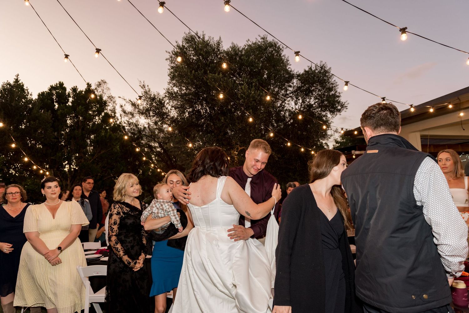 Guests join bride and groom on dance floor at outdoor wedding reception under string lights.