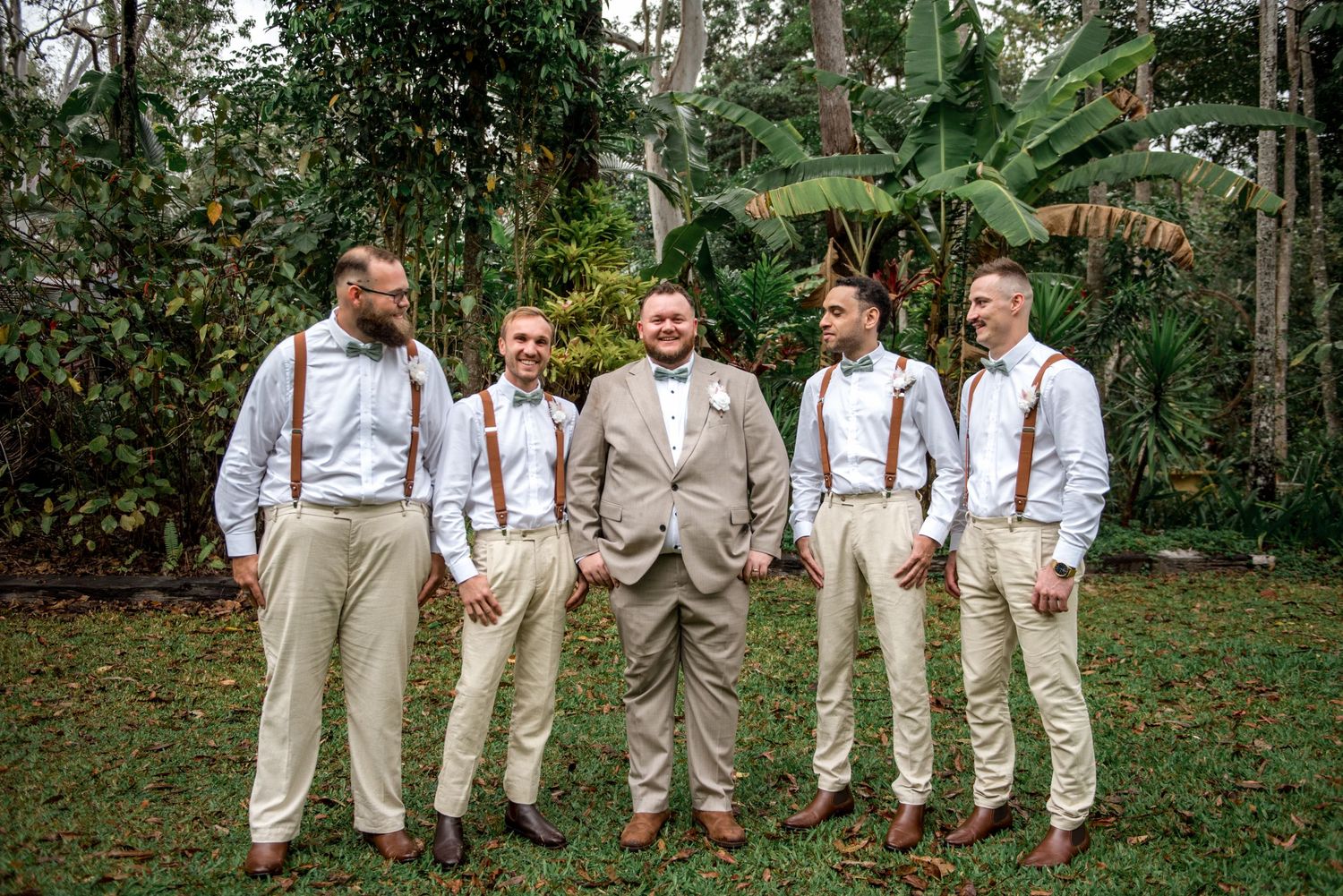 Groom in tan suit stands with four groomsmen in white shirts and brown suspenders in tropical garden.