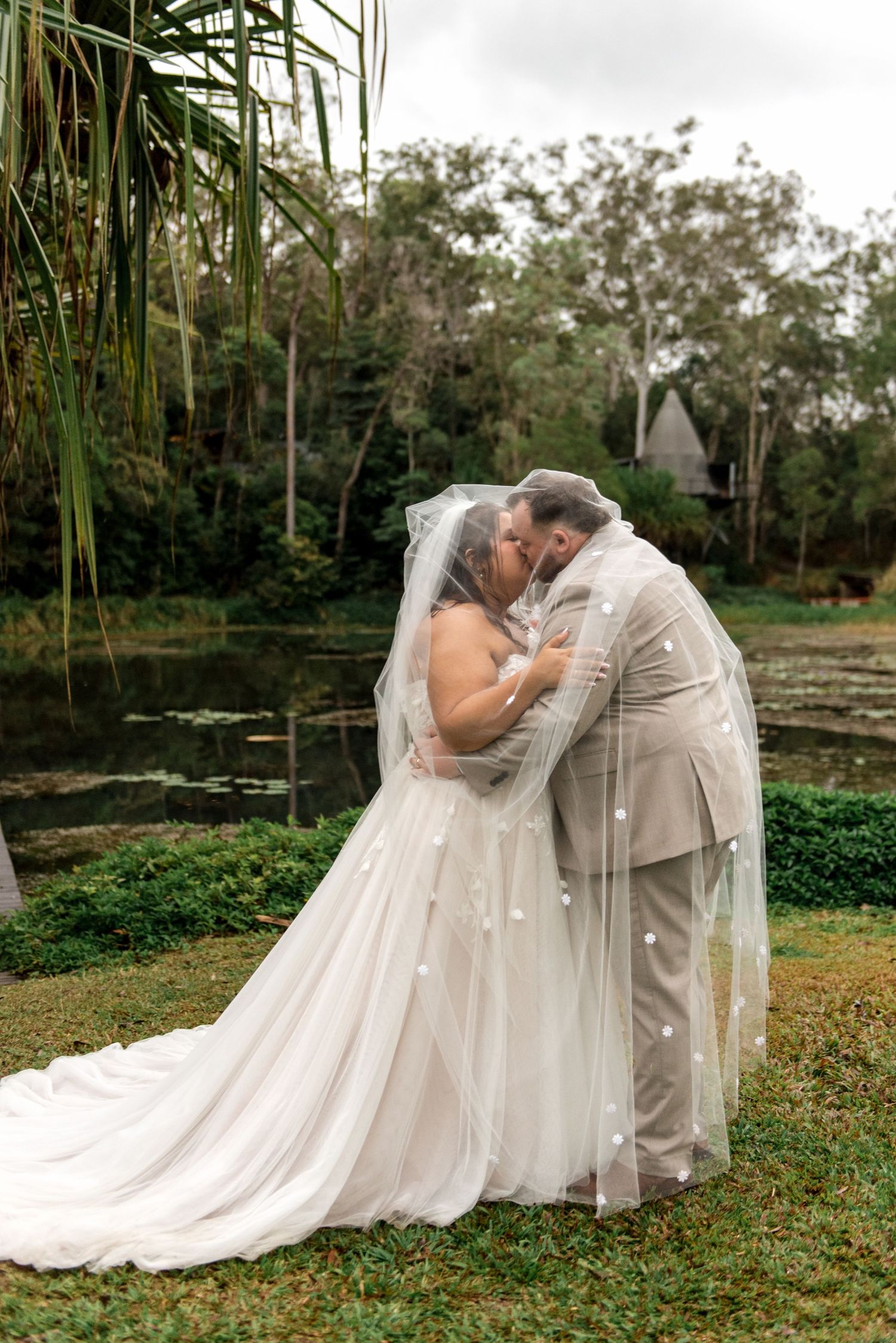 Bride and groom share a romantic kiss under a pearl veil near a serene lily pond.